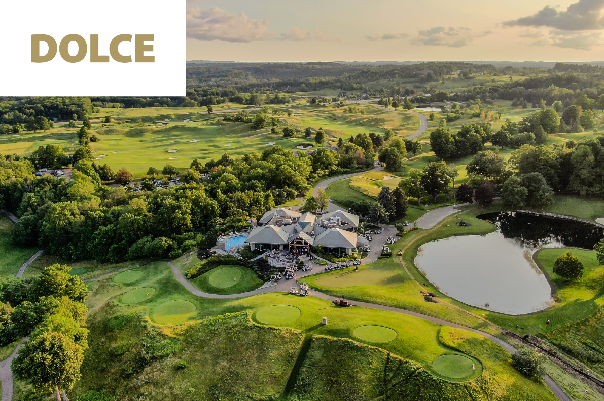 Aerial view of a golf course with green fairways, sand traps, a clubhouse, a pond, trees, and surrounding landscape during daytime.