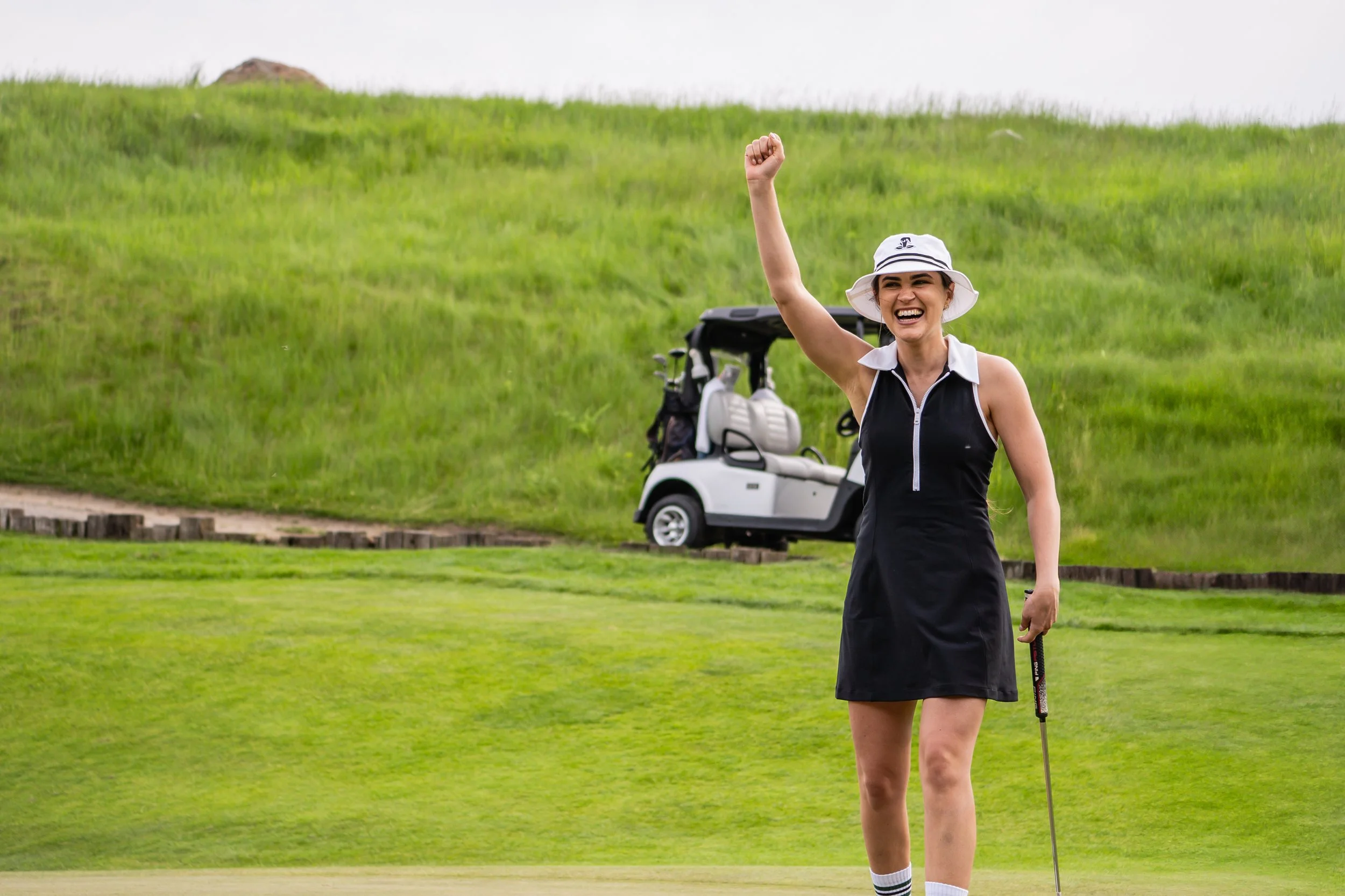 A woman in a black and white golf outfit with a white hat, smiling and raising her fist in celebration on a golf course, with a golf cart and green hills in the background.