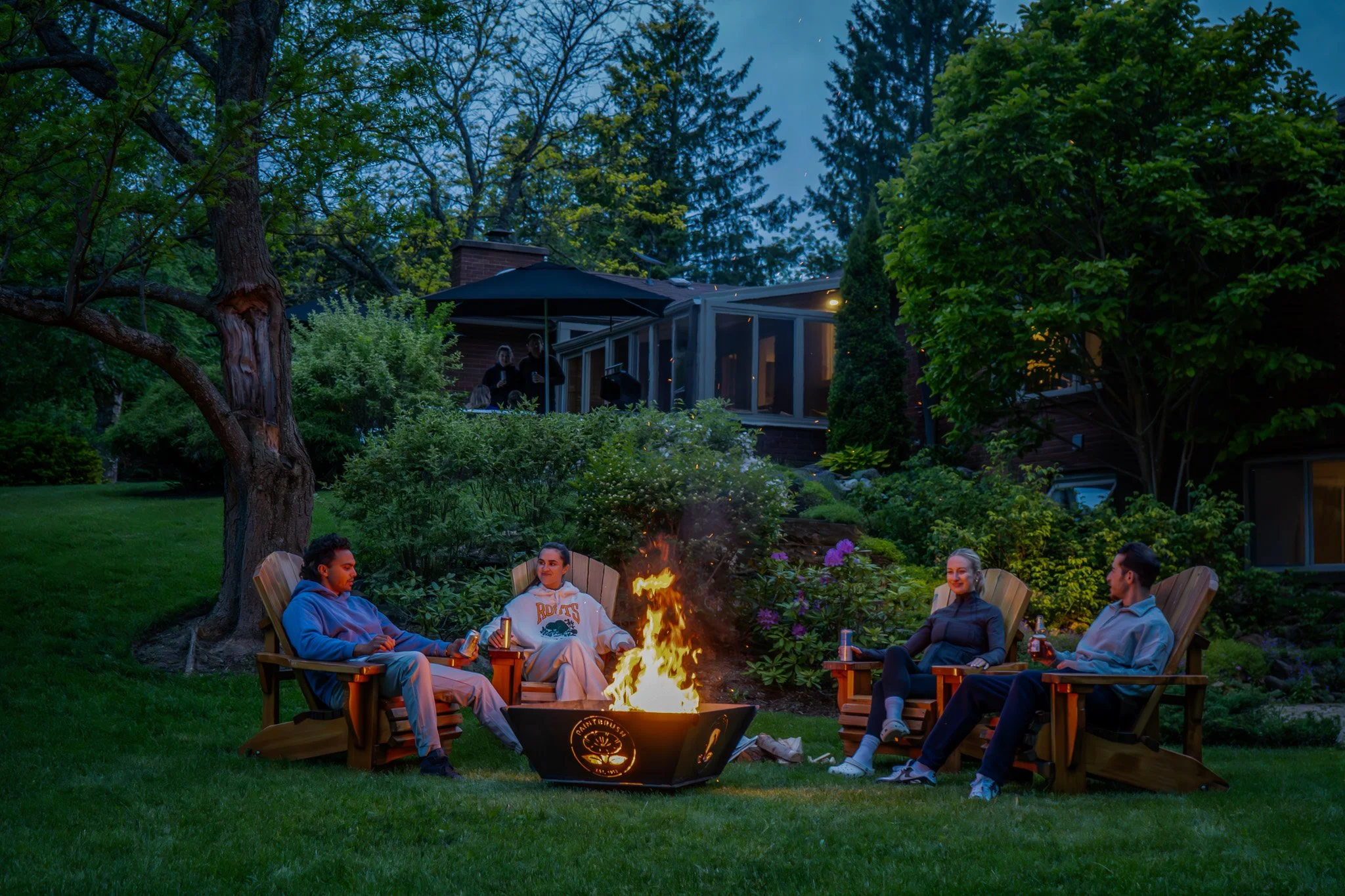 Four people sitting around a fire pit on a lawn at dusk, with a house and trees in the background.