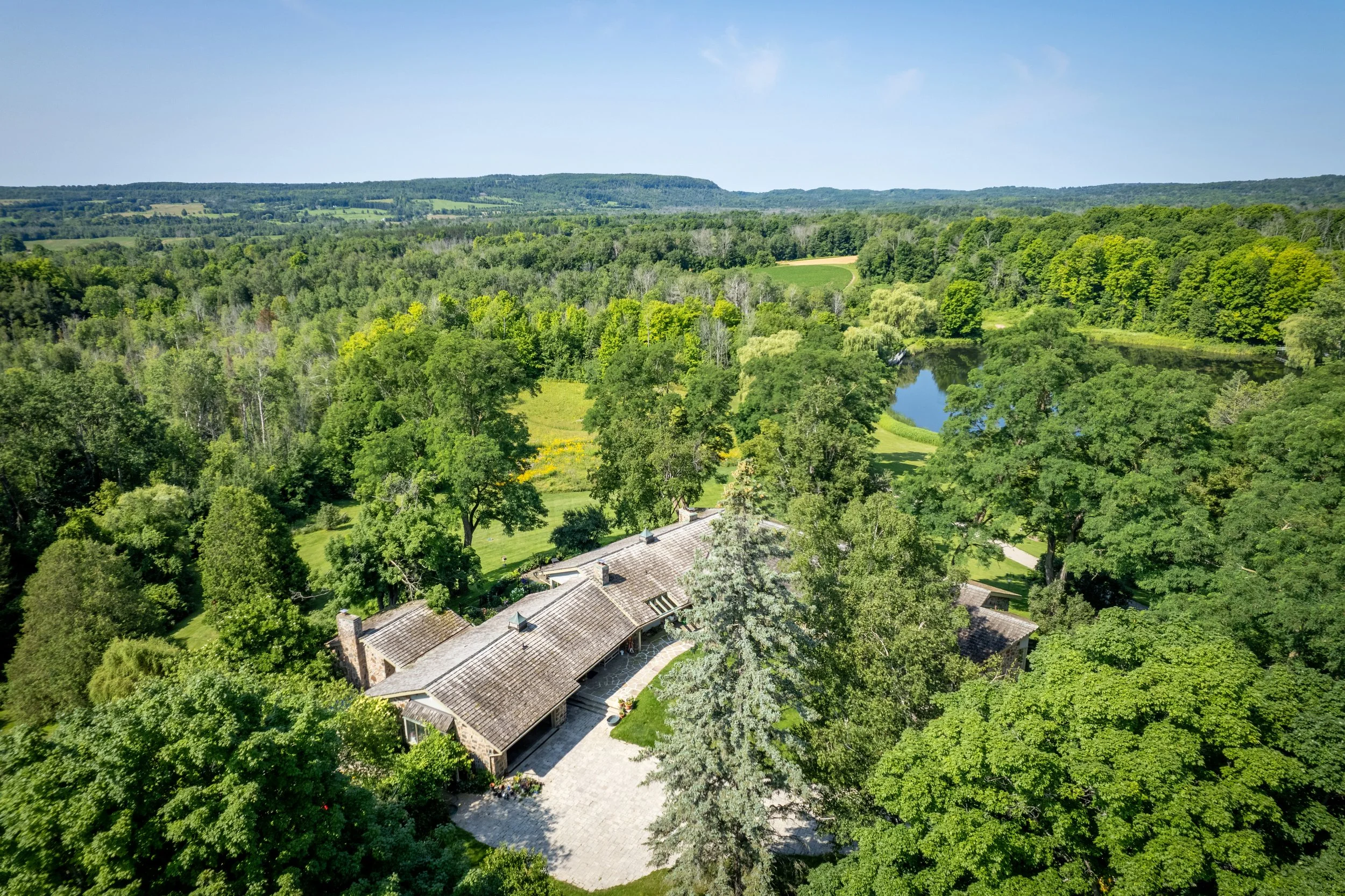 Aerial view of a large house surrounded by lush green trees, with a small pond and open fields in the background under a clear blue sky.
