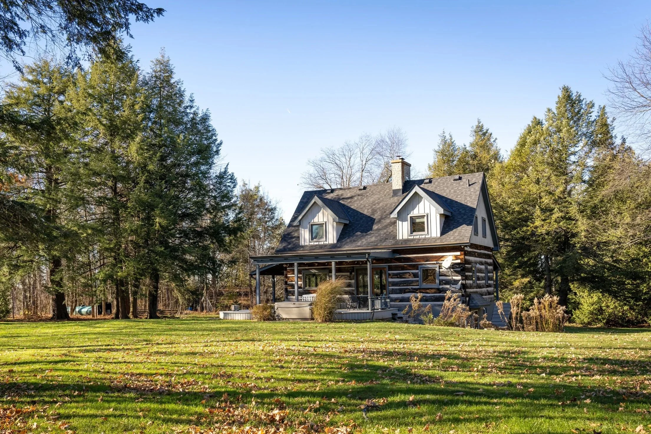 A rustic two-story house with a porch, set in a lush yard with large trees and fallen autumn leaves, under a clear blue sky.