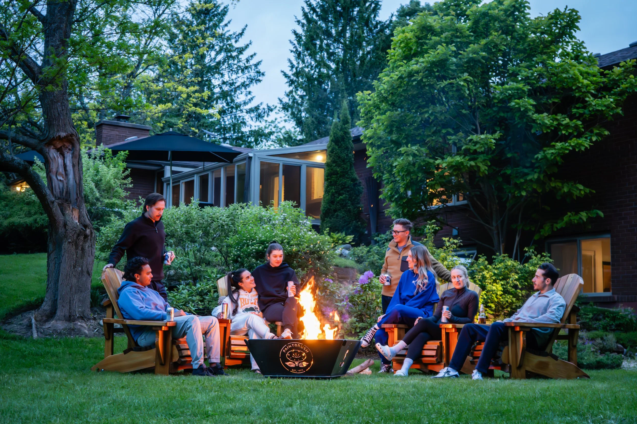 Group of friends gathered around a campfire in a backyard with trees and a house in the background during evening.