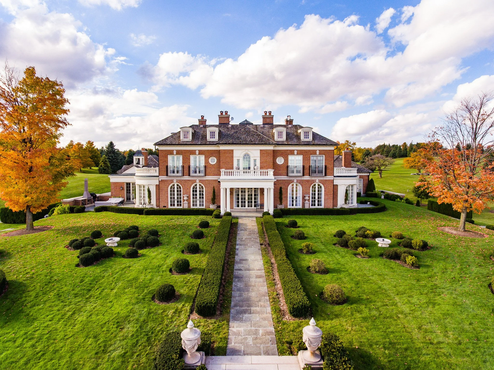 Large brick mansion with white accents, multiple balconies, and a long stone pathway leading to the entrance, surrounded by well-manicured lawn and trees with autumn foliage.