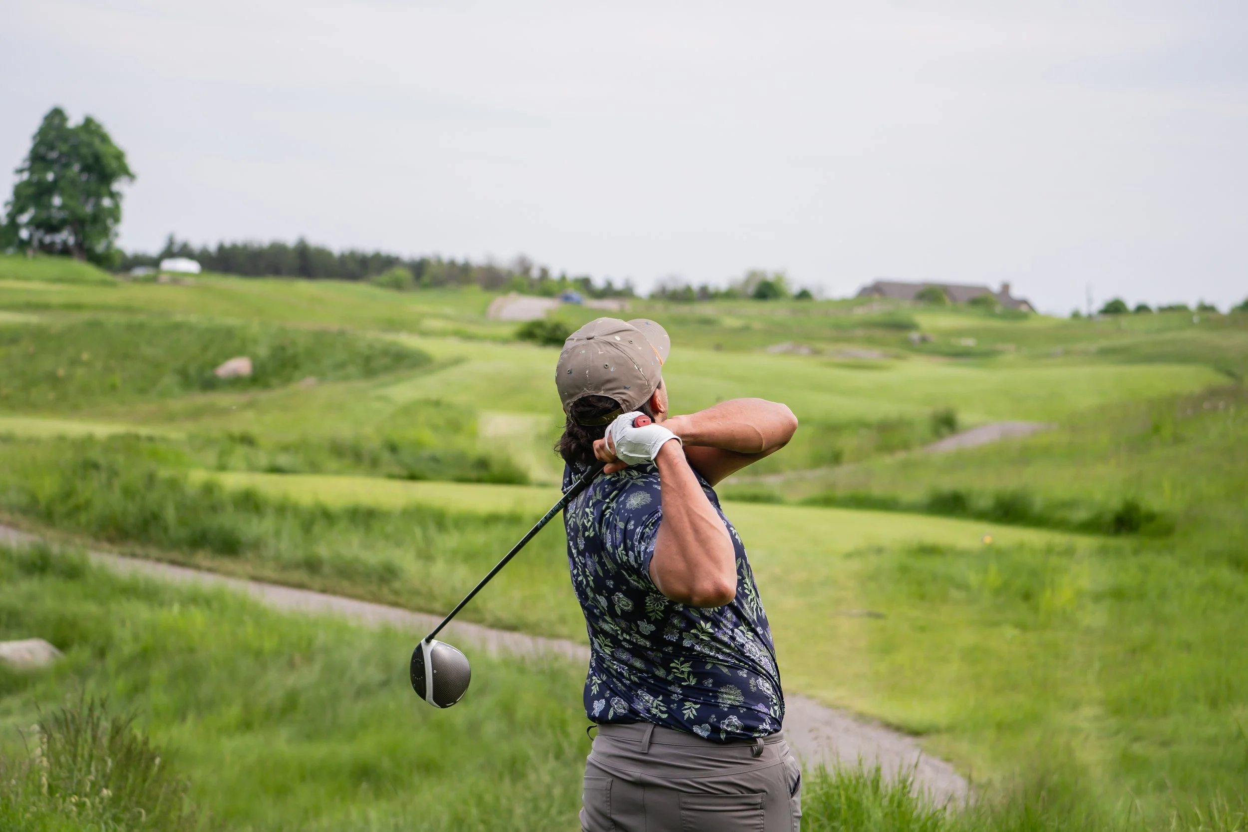 A person golfing on a lush green golf course with rolling hills in the background, holding a golf club over their shoulder and watching where the ball goes.