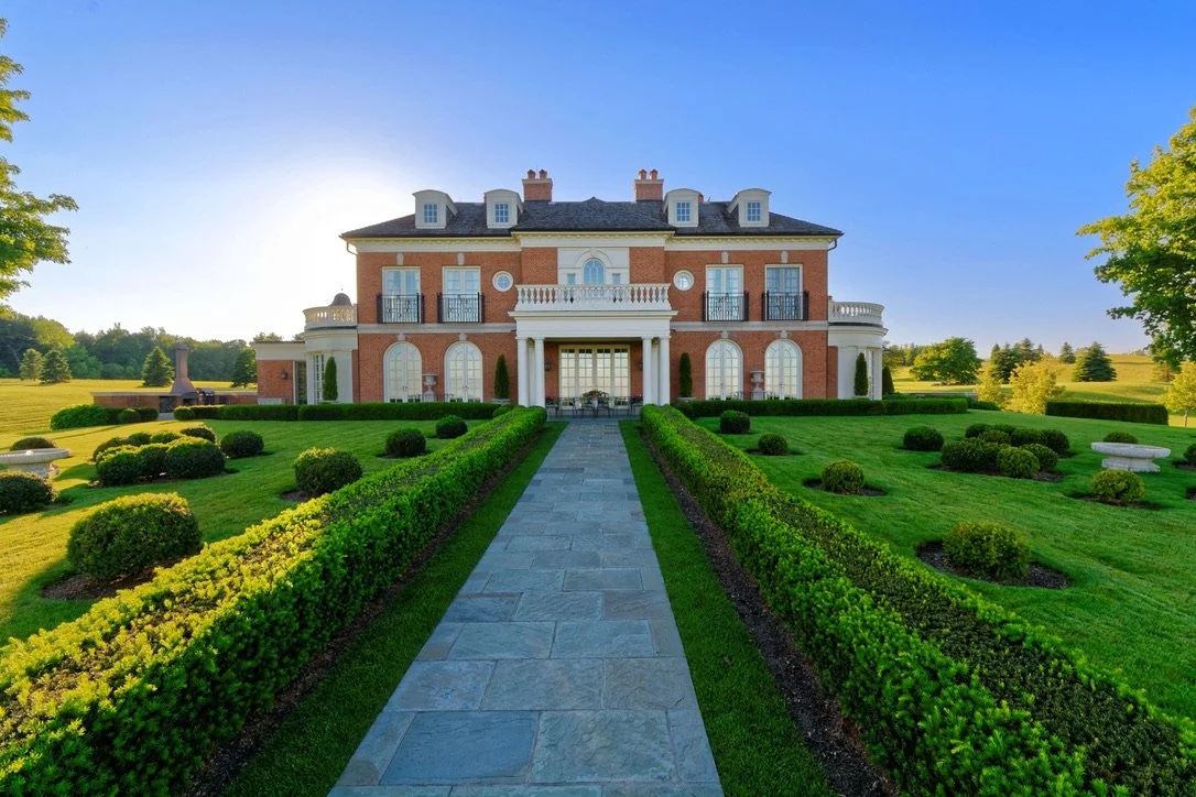 Front view of a large, elegant brick mansion with a paved walkway leading to the entrance, surrounded by well-manicured green lawns and shrubs, with trees and open fields in the background under a clear blue sky.