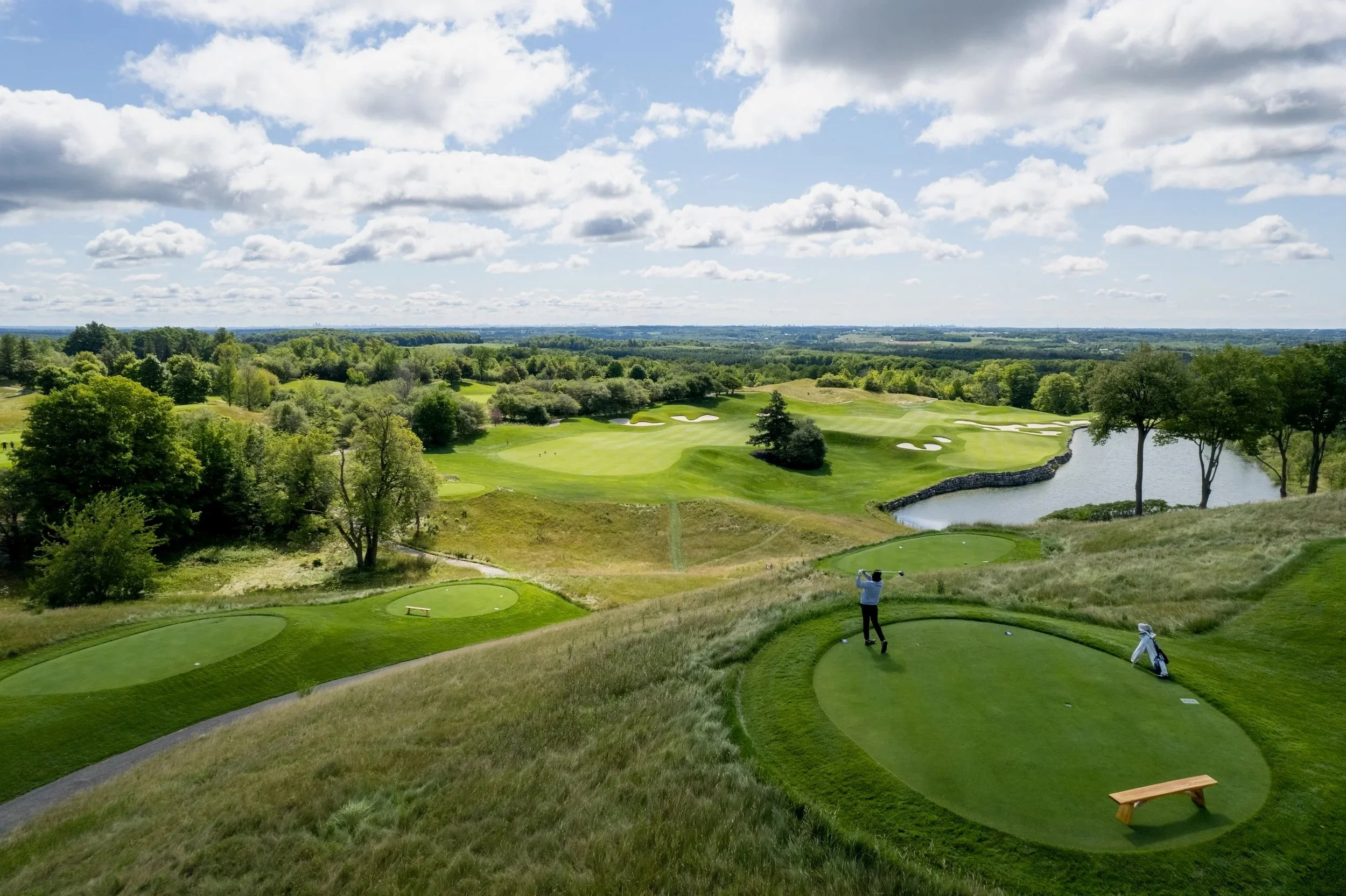 A scenic golf course with green fairways, sand bunkers, a water hazard, and a golfer in mid-swing on the tee box, surrounded by trees and a partly cloudy sky.