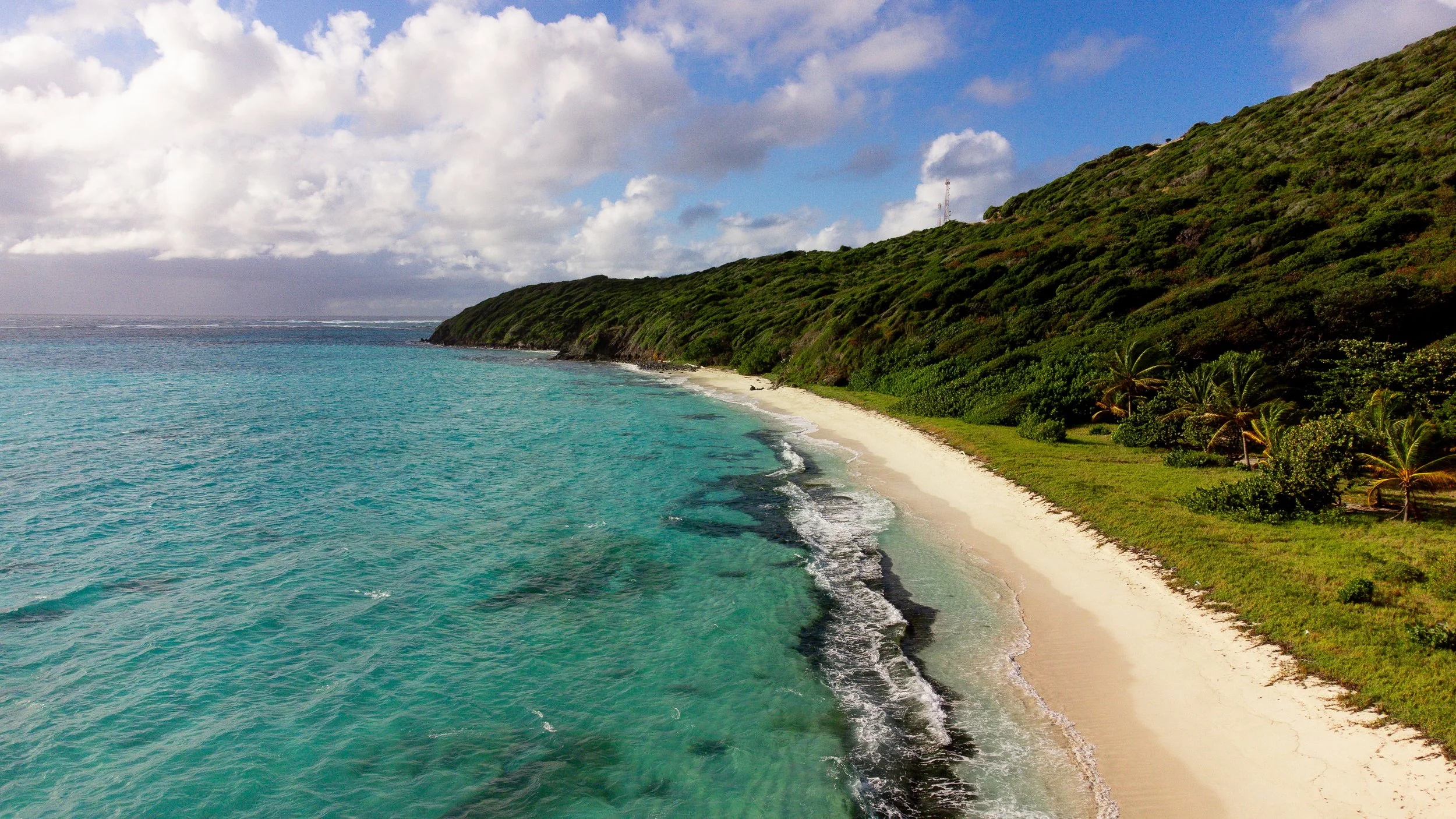 A tropical beach with clear turquoise water, white sandy shoreline, and green hills with palm trees under a partly cloudy sky.