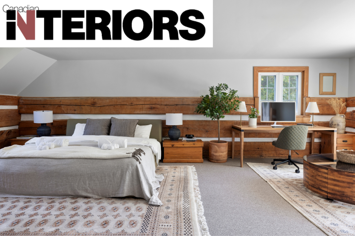 A bedroom featuring a bed with a beige bedspread, multiple pillows, and rolled towels, wooden nightstands with lamps, a potted plant, a desk with a computer monitor, and a window with natural light.