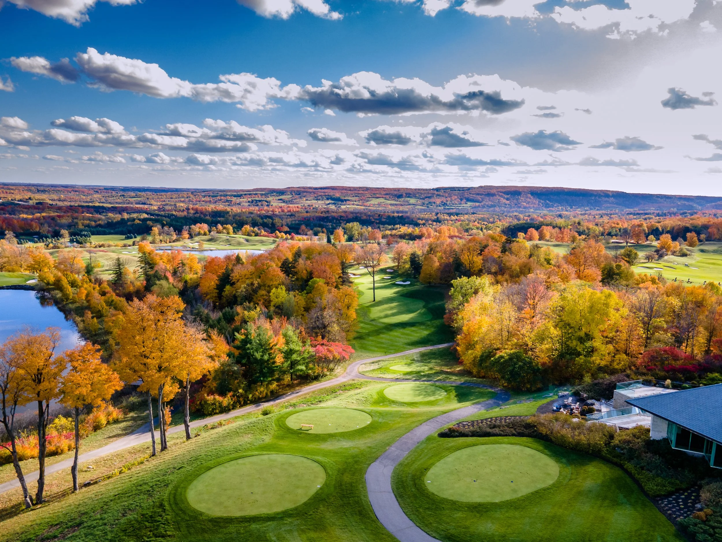 An aerial view of a golf course surrounded by colorful autumn trees, with a lake on the left and a clubhouse on the bottom right, under a partly cloudy sky.
