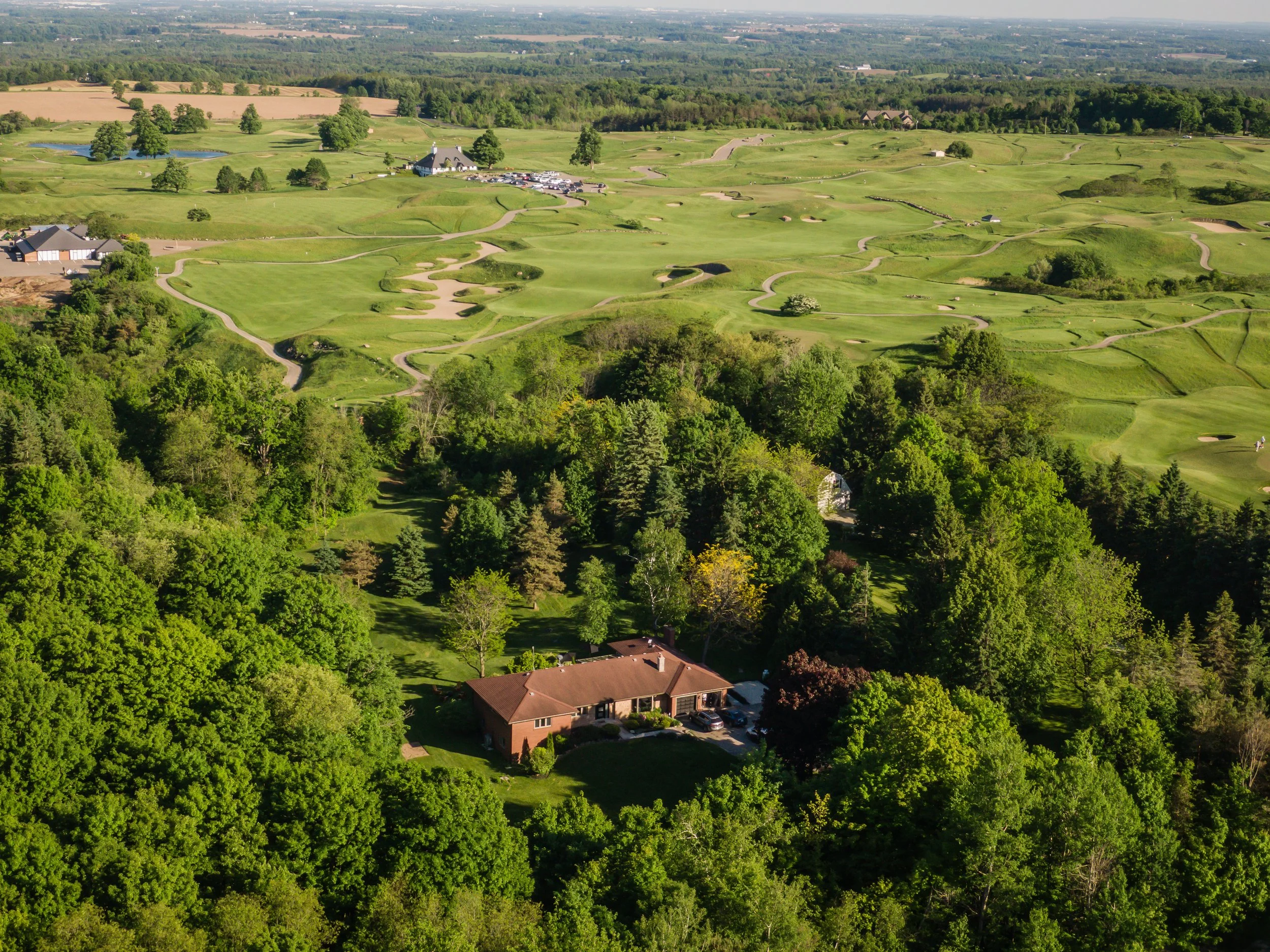 Aerial view of a golf course with green fairways, sand traps, and trees, along with houses in the foreground surrounded by lush greenery.