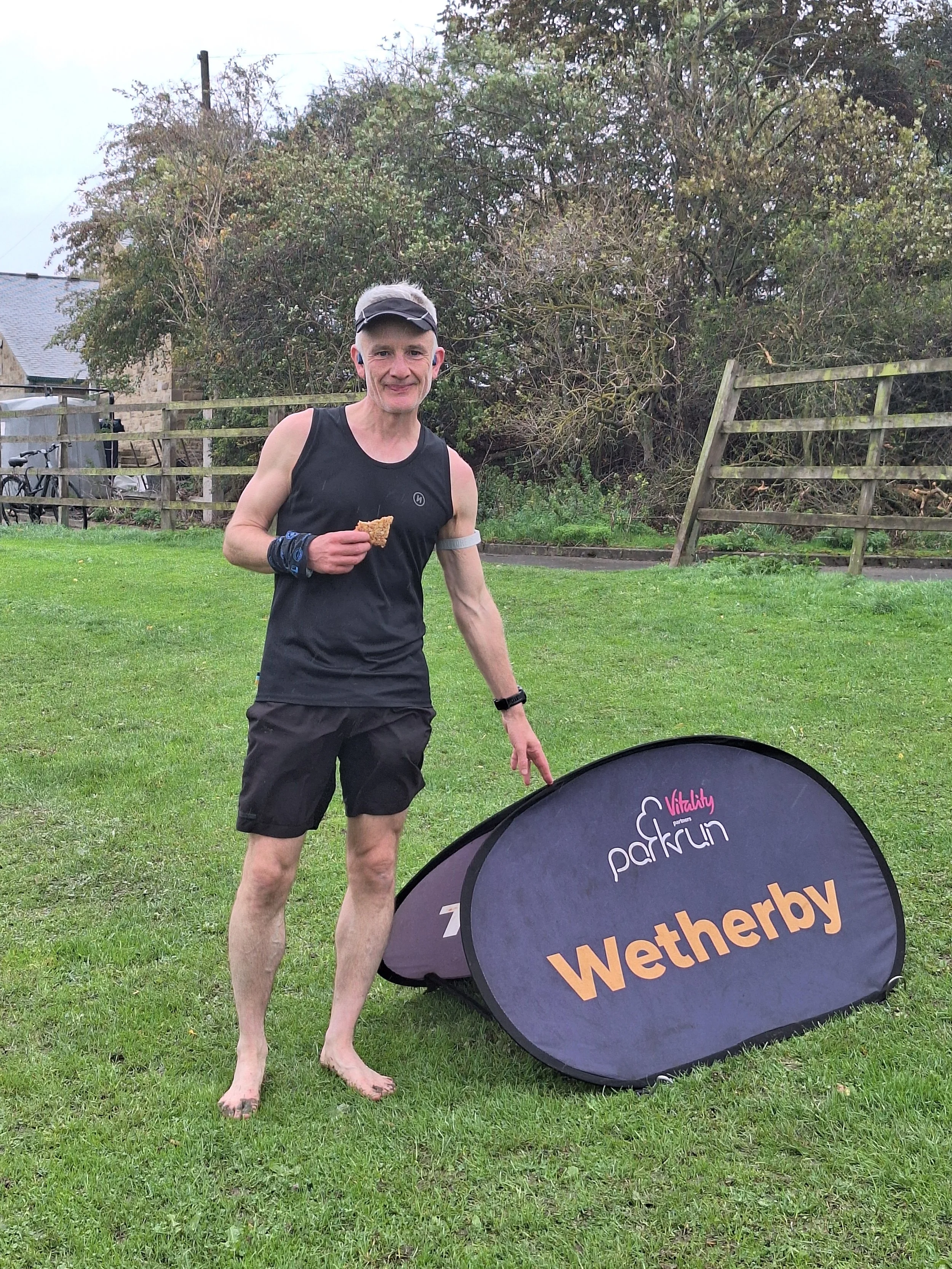 A man in running gear, holding a snack, standing next to a Wetherby banner at a park or race event.