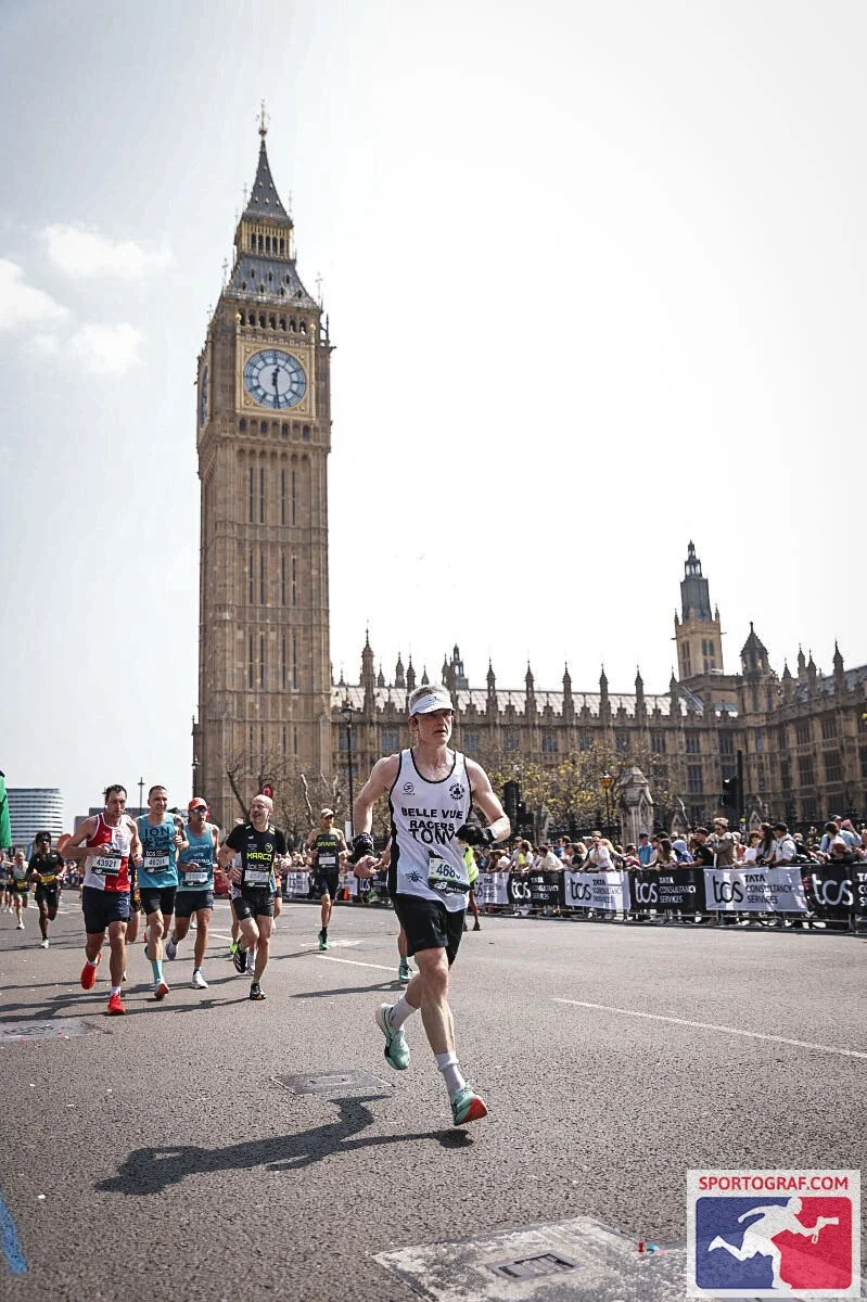Marathon runners running in front of Big Ben in London, with spectators watching behind barriers and an overcast sky.