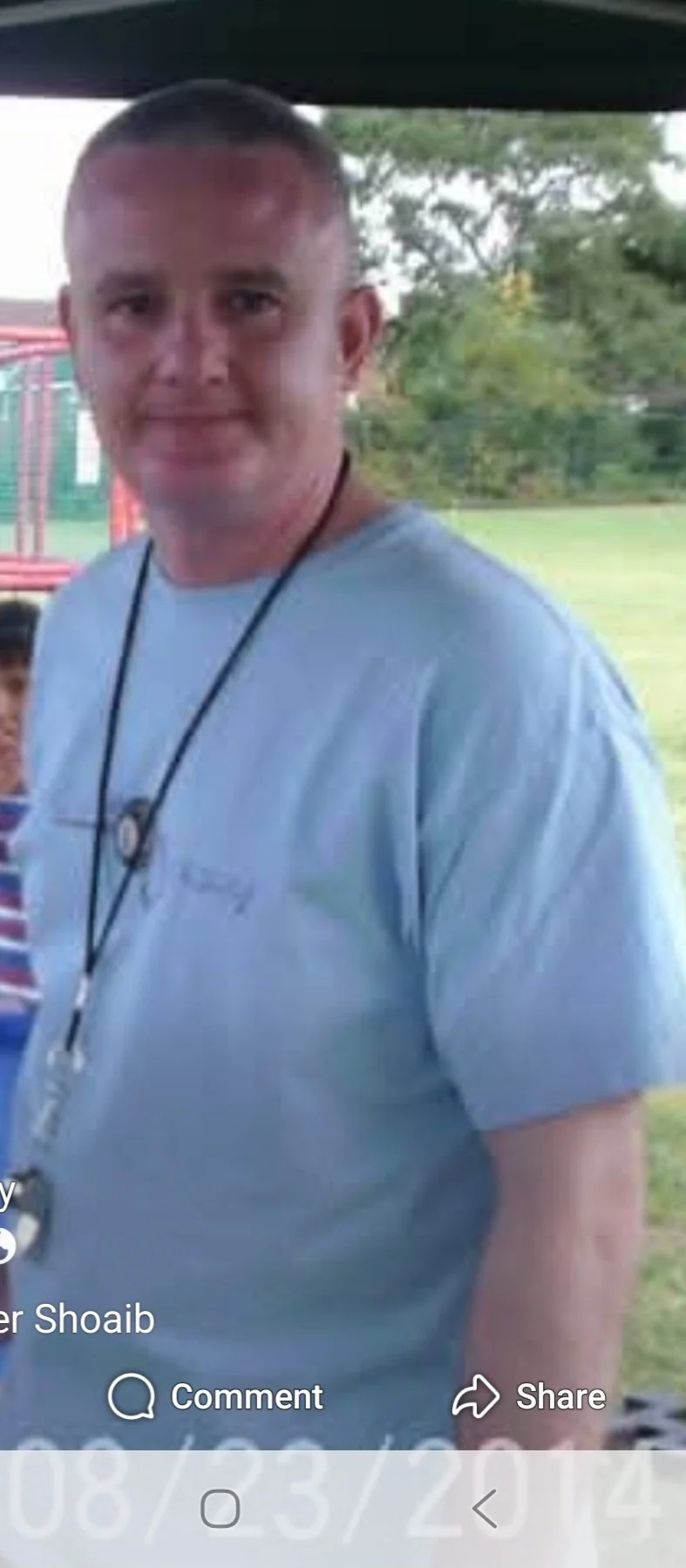 A man with short hair and a light blue t-shirt standing outdoors near playground equipment with trees in the background.