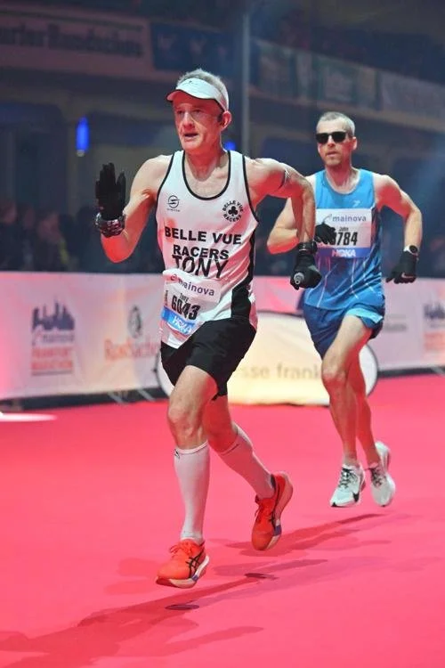 Two marathon runners compete on a pink track with banners and spectators in the background. The runner in front is wearing a white cap, arm warmers, black shorts, and a white tank top with "BELLE VUE RACERS TONY" written on it, and orange running shoes. The runner behind is in sunglasses, a blue tank top, black shorts, and gray running shoes.
