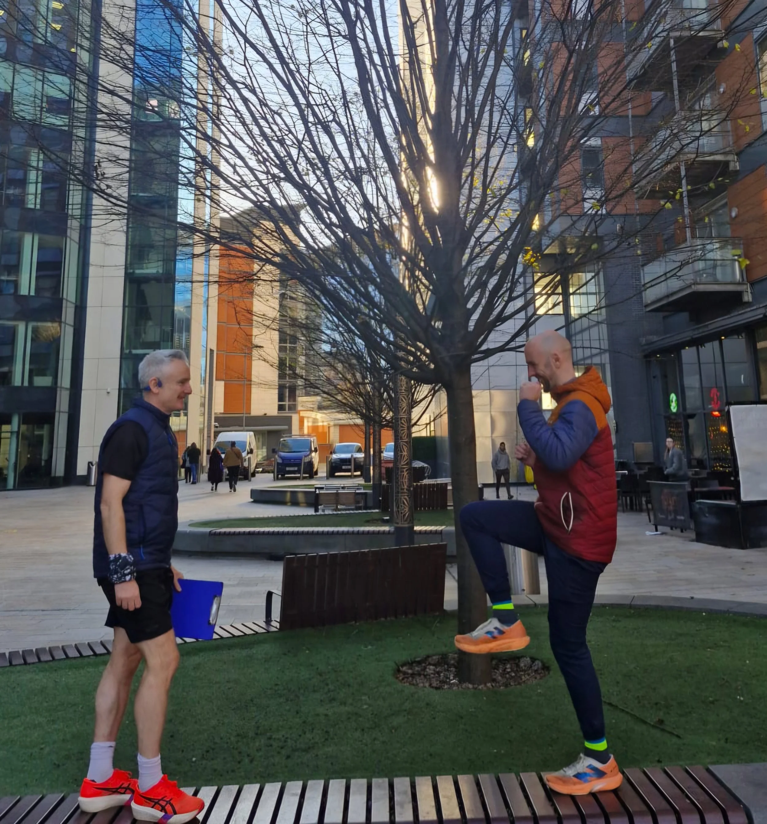 Two men are outdoors in a city park, smiling and laughing. One man is standing and the other is lifting his knee as if in a playful manner. There is a leafless tree in the background and modern buildings surrounding the park.