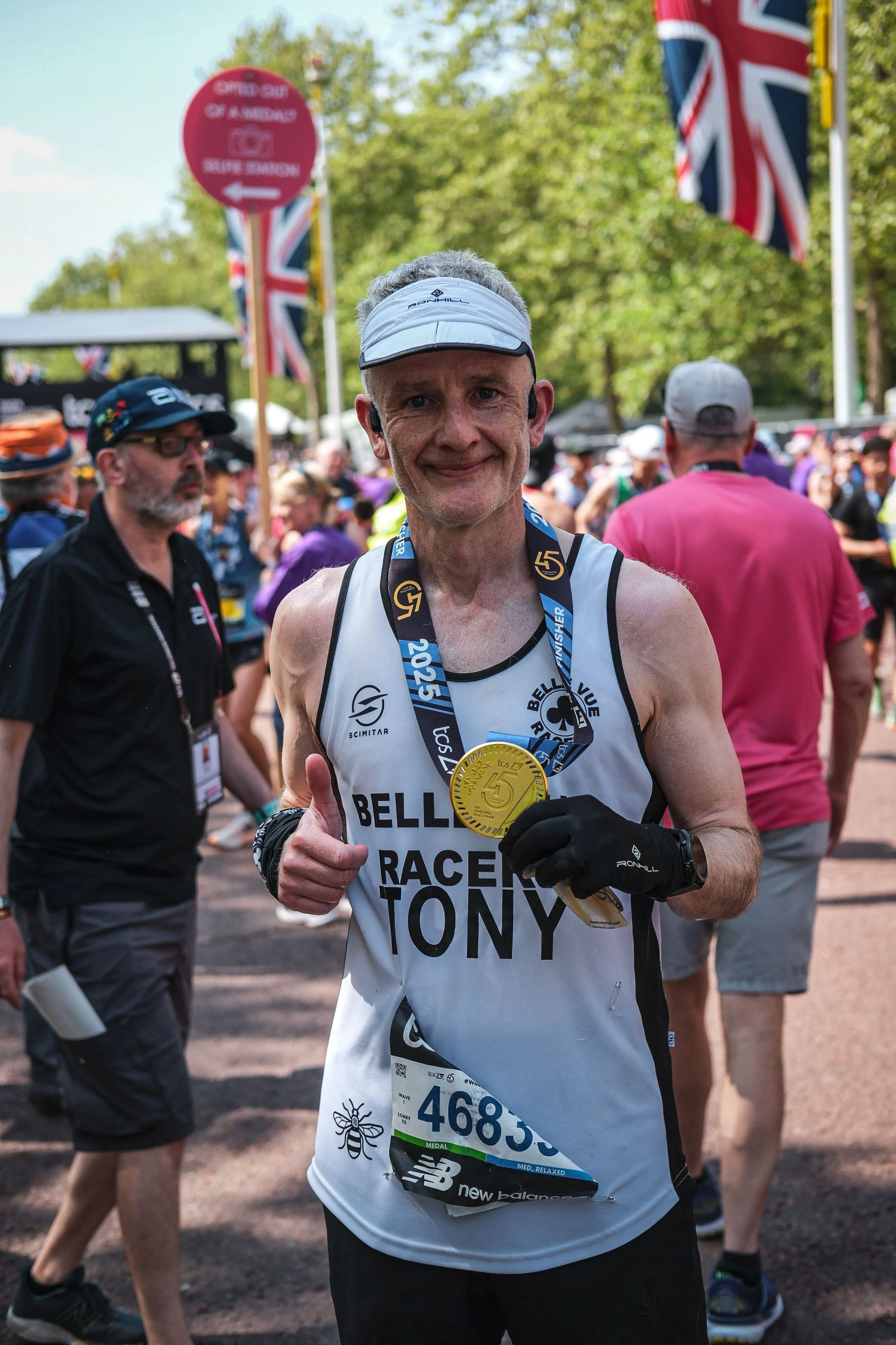 A man in marathon running gear, wearing a white visor and a Medal of the 2023 London Marathon, smiling and giving a thumbs-up at the finish line surrounded by other runners and spectators.