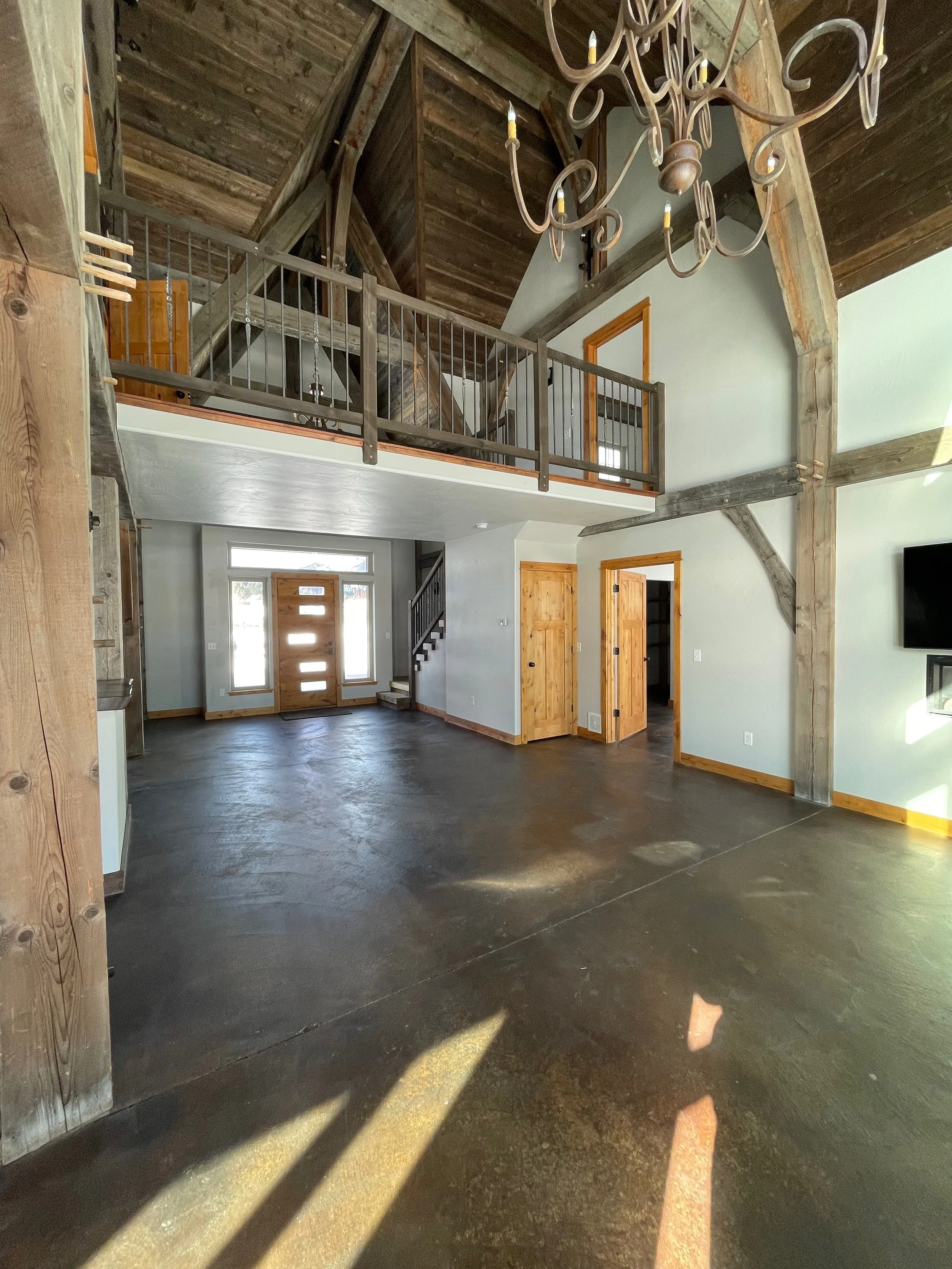 Interior of a spacious, rustic-style home with high vaulted ceilings, exposed wooden beams, and a polished concrete floor, featuring a chandelier and an open entryway with a front door and staircase.
