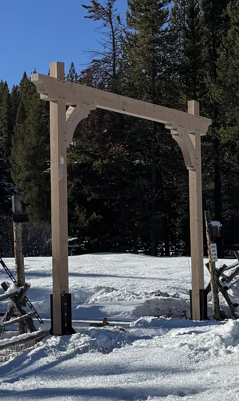Wooden archway construction in snowy outdoor setting with evergreen trees in the background.
