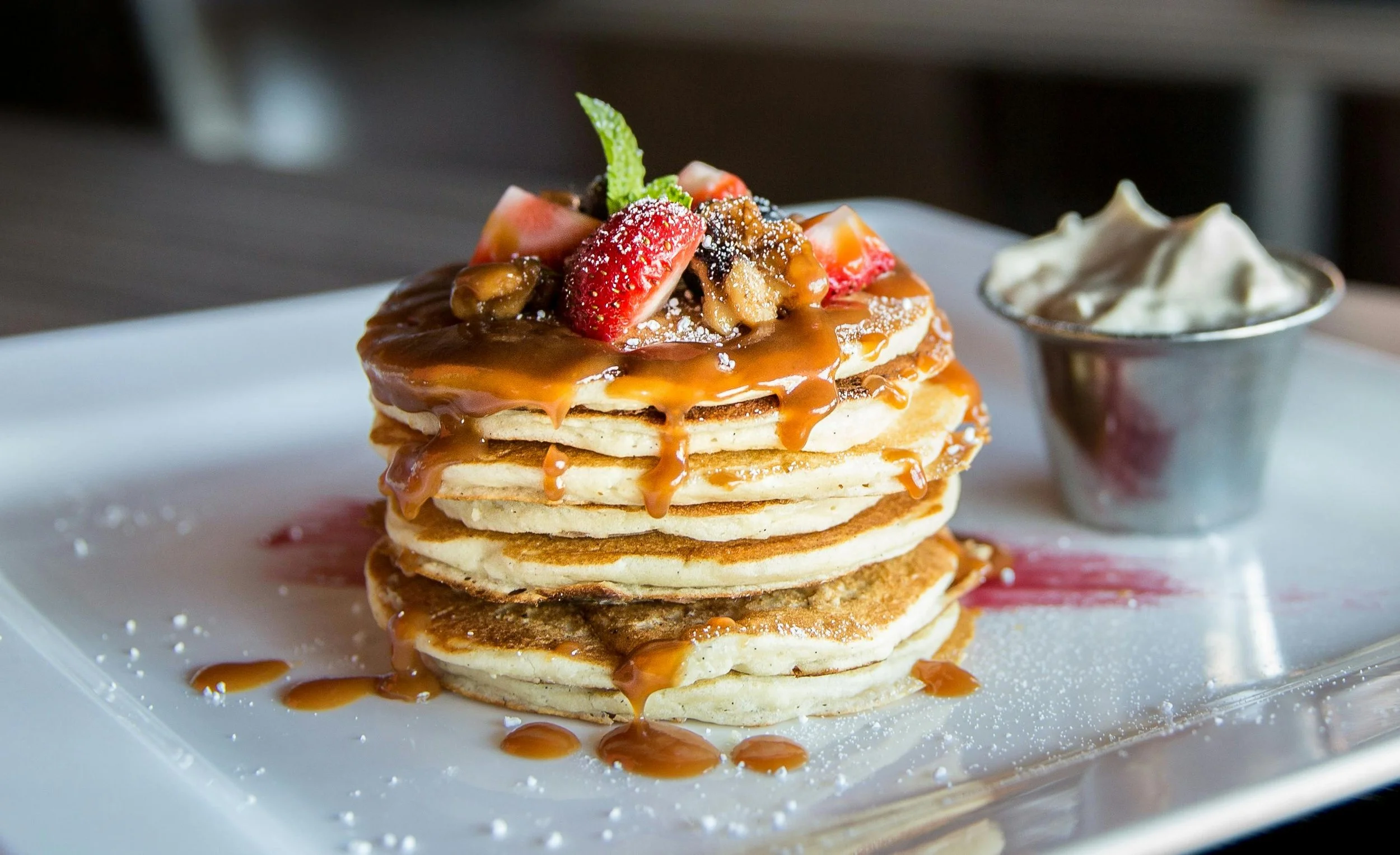 Stack of four pancakes topped with strawberries, caramel, and powdered sugar, served with whipped cream on the side.