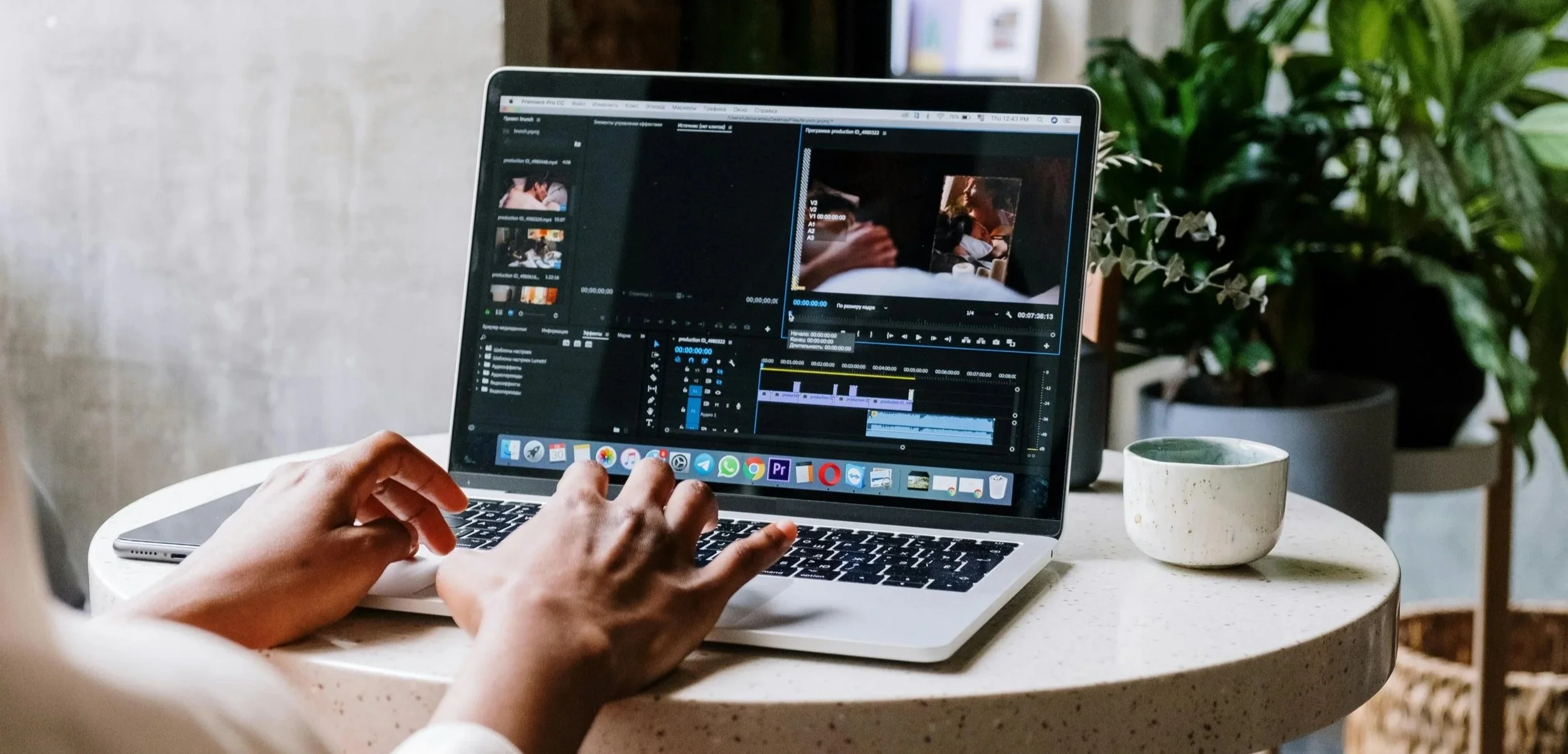 Person editing a video project on a MacBook laptop, placed on a round table with a white speckled surface, next to a ceramic coffee mug, with potted plants in the background.