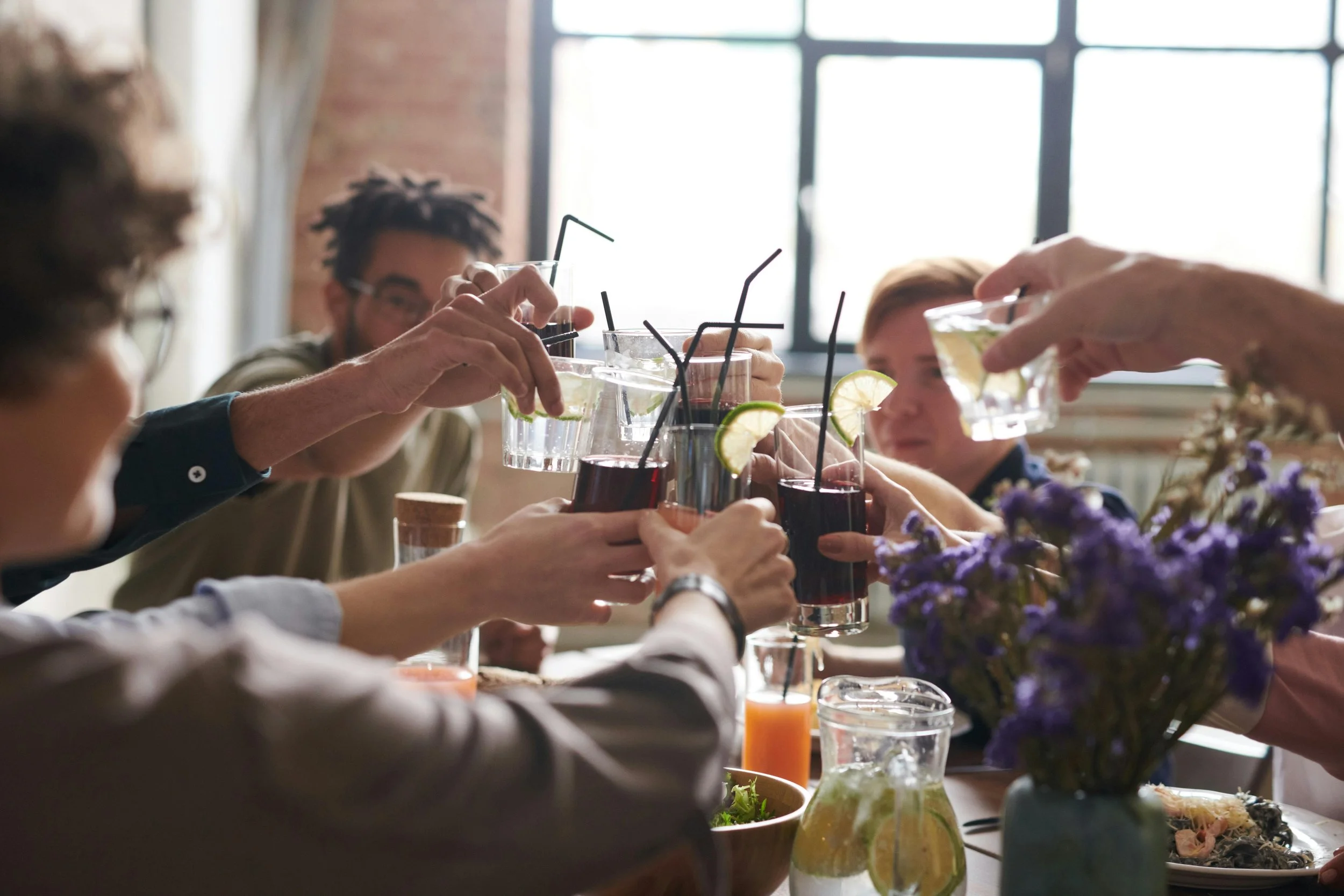 People raising glasses for a toast at a restaurant or gathering, with drinks garnished with lemon and lime, a vase with purple flowers, and a window in the background.