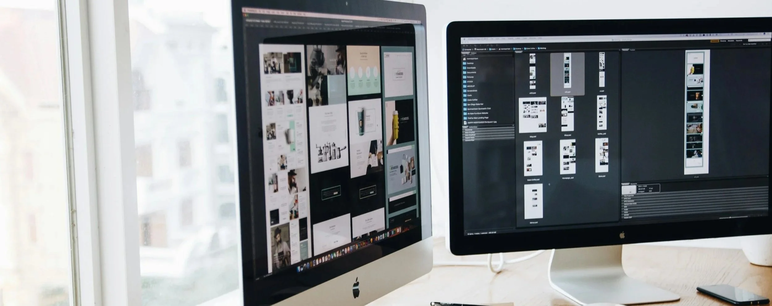 Two Apple computer monitors on a wooden desk, displaying digital design and photo editing software. Bright light coming through a nearby window.