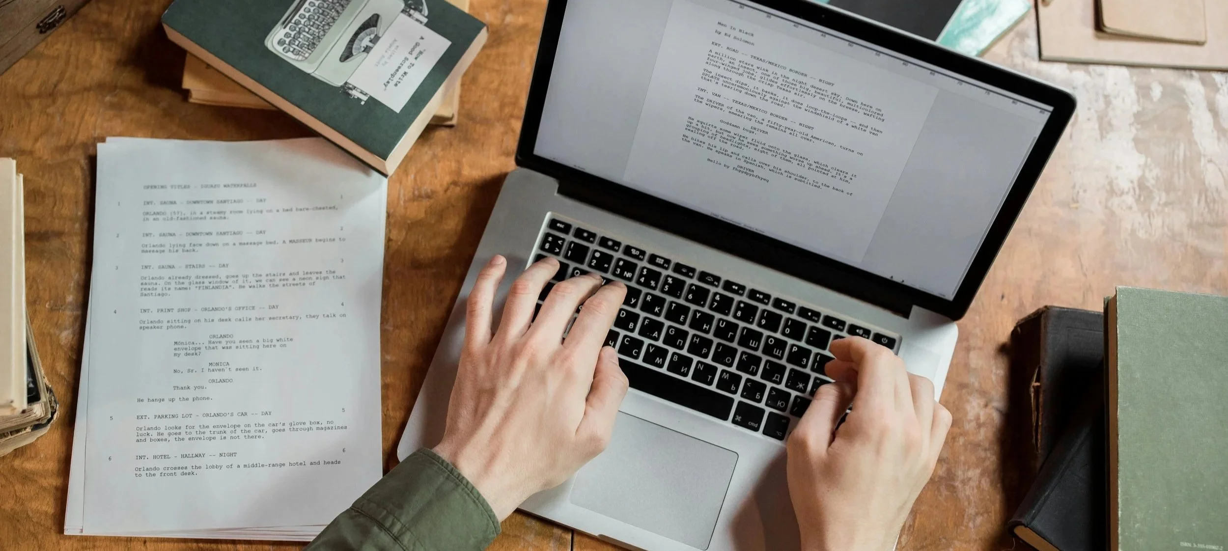 A person using a laptop on a wooden desk, with sheets of paper, books, and other documents scattered around.