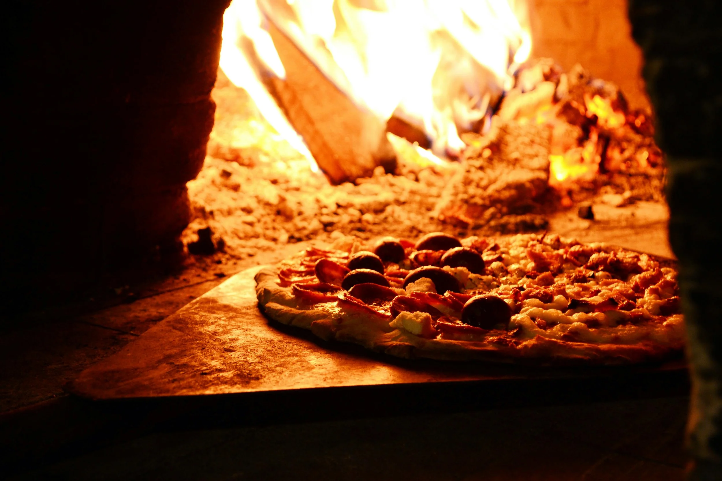 Pizza being baked in a wood-fired oven with flames visible in the background.