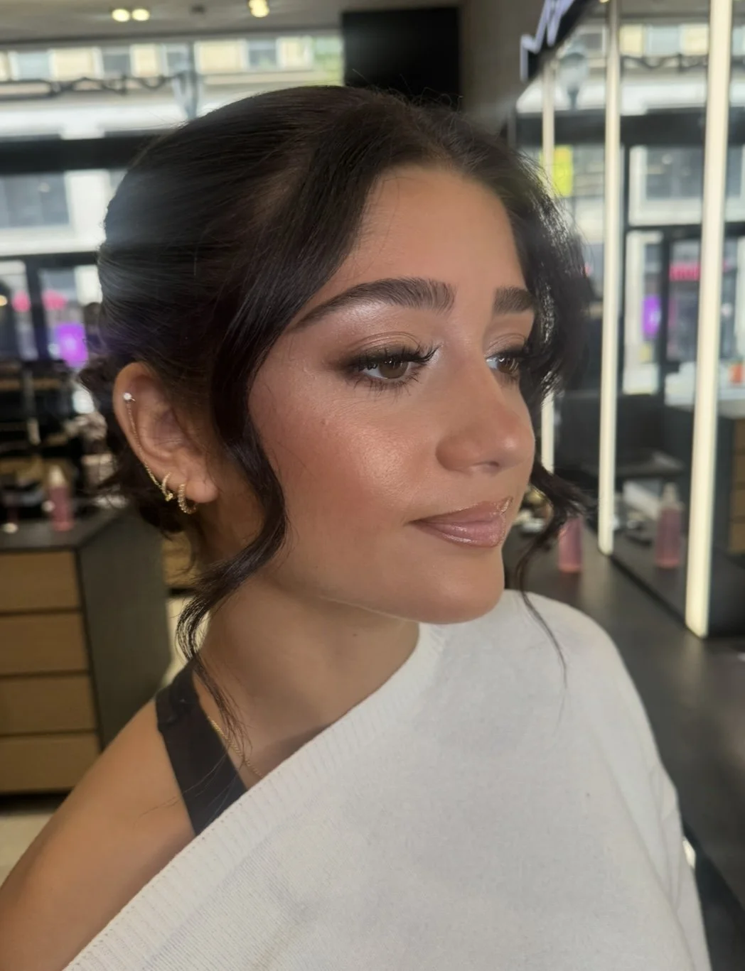 Close-up of a woman with dark wavy hair, makeup, and jewelry, looking to the side in an indoor setting with shelves and windows in the background.