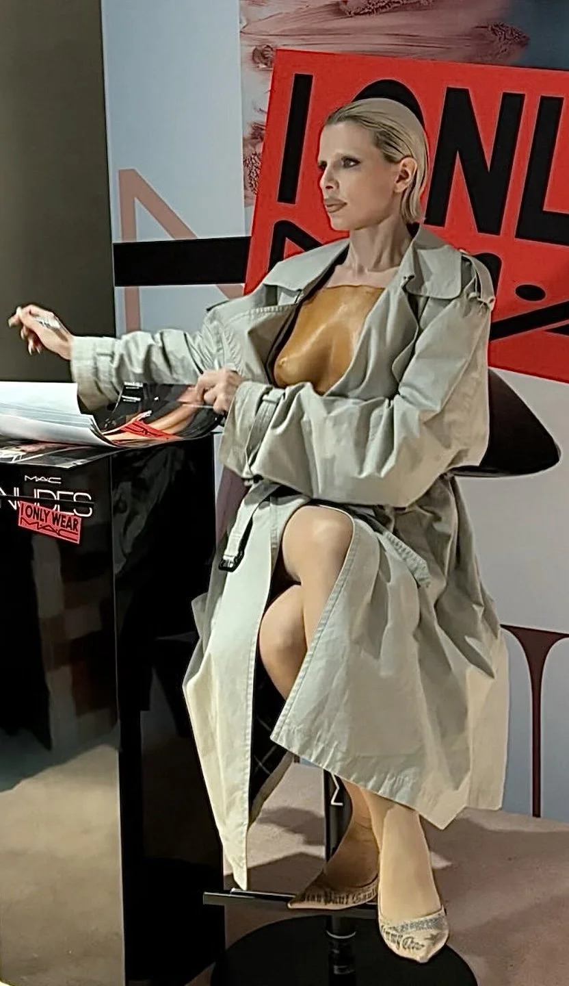A woman with blonde hair in a trench coat and heels, sitting at a table with a Mac cosmetics box and promotional materials, with an 'ICON' sign and protest signs in the background.