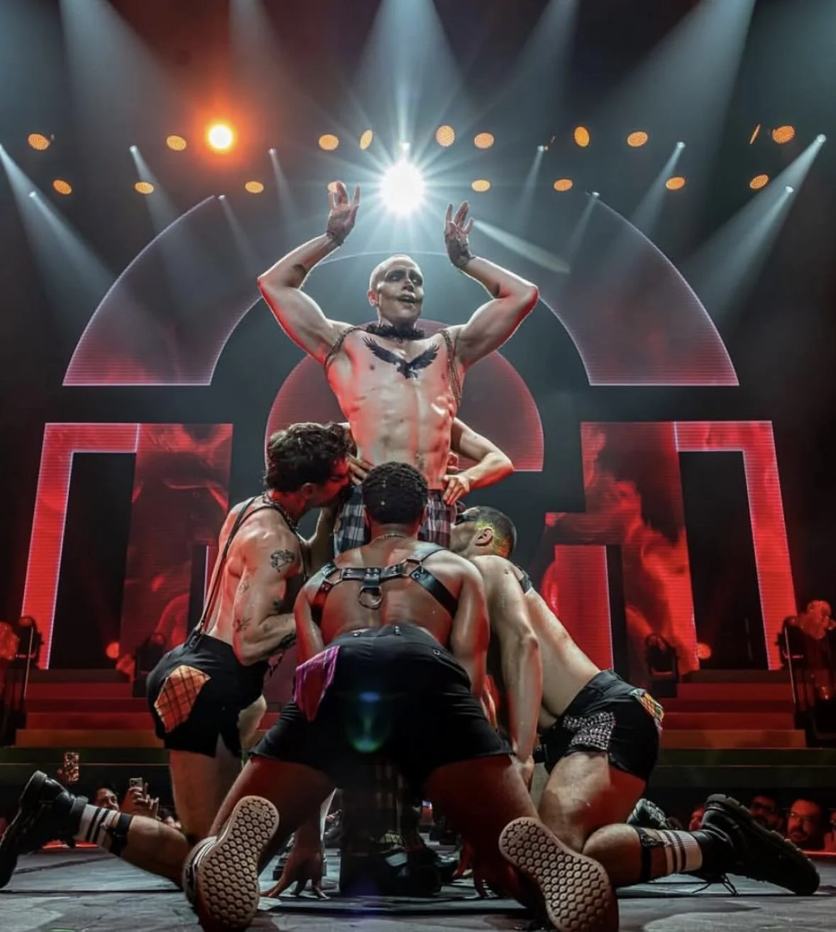 Performer on stage with five men kneeling and supporting him, stage lighting and digital screen in the background.