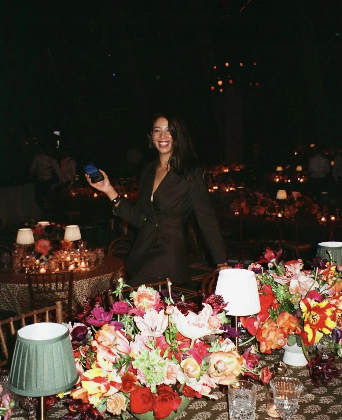 A woman in a black dress smiling and holding a phone at an elegant dinner event with floral centerpieces and dim lighting.