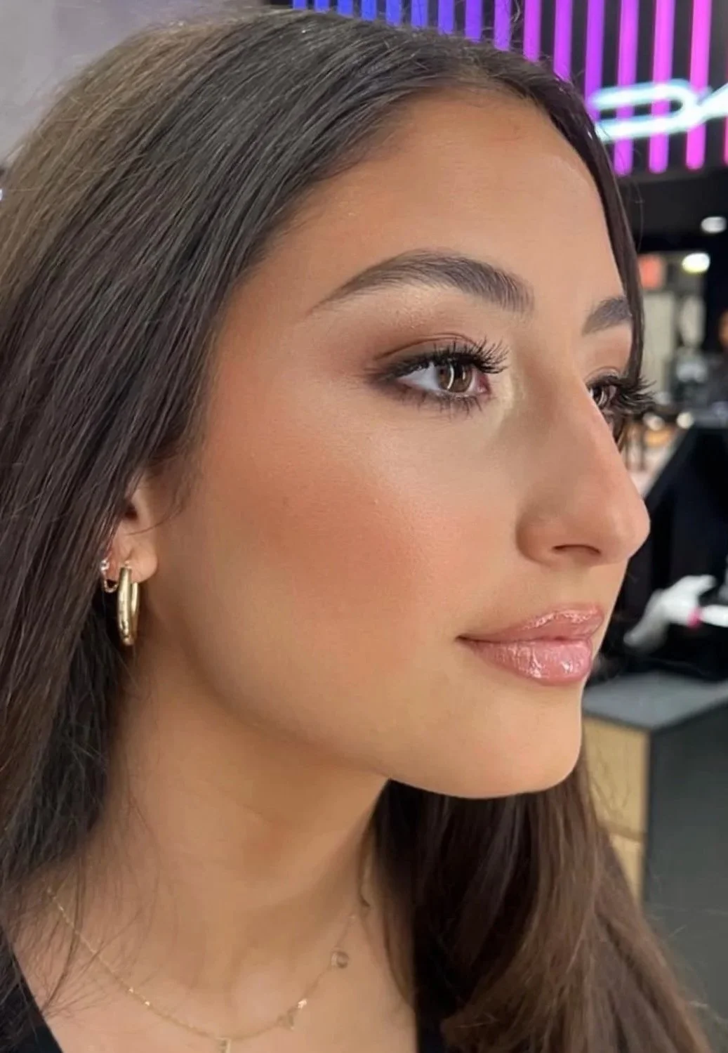 A close-up side profile of a woman with long brown hair, wearing gold hoop earrings and layered necklaces, with makeup including nude lipstick, blush, eye shadow, and mascara, in an indoor setting with neon purple and blue lighting in the background.