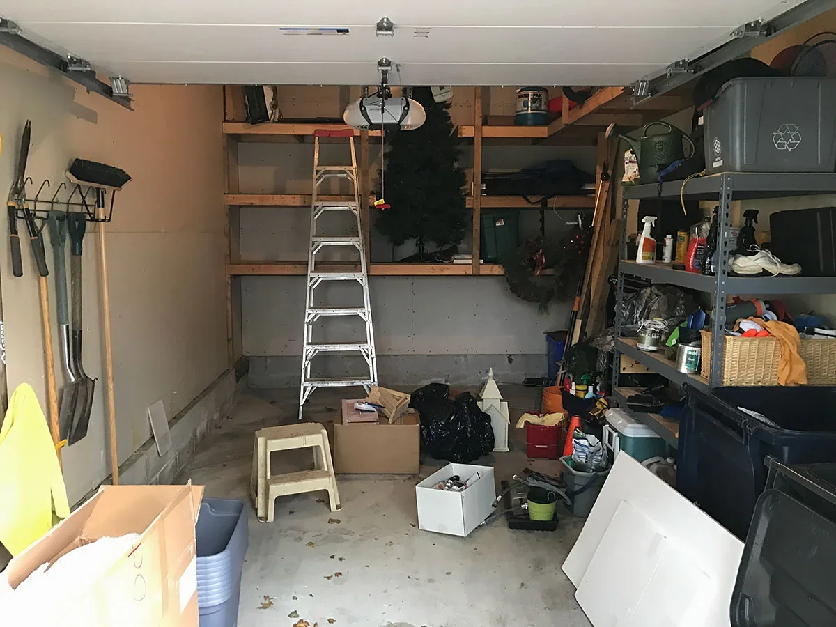 A cluttered garage with tools hanging on the wall, a ladder, storage shelves filled with various items, and miscellaneous clutter on the floor.