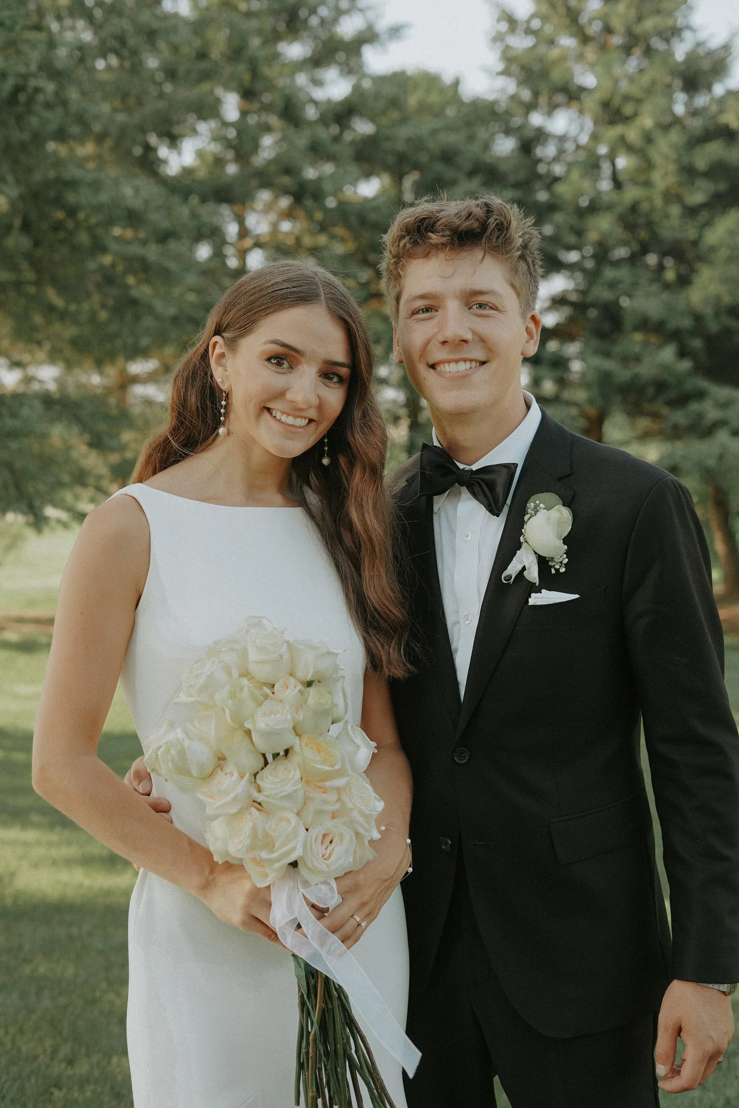 Bride and groom standing outdoors, smiling for a wedding photo, with trees in the background. The bride holds a bouquet of white roses, wearing a sleeveless white wedding dress and pearl earrings. The groom wears a black tuxedo with a bow tie and boutonniere.