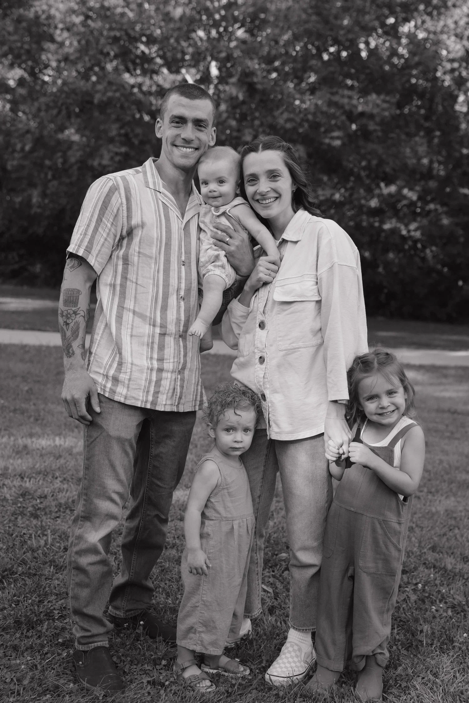 A black and white photo of a family of six standing outdoors on grass, smiling at the camera. The family includes two adults, one man and one woman, and four young children.