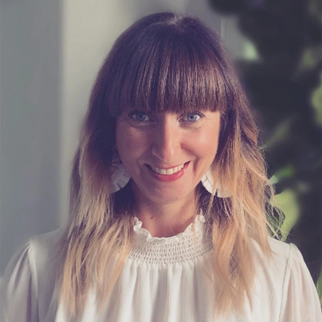 A woman with shoulder-length light brown hair, wearing a white top with ruffled neckline, smiling with visible teeth, and blue eyes. Blurred background with greenery.