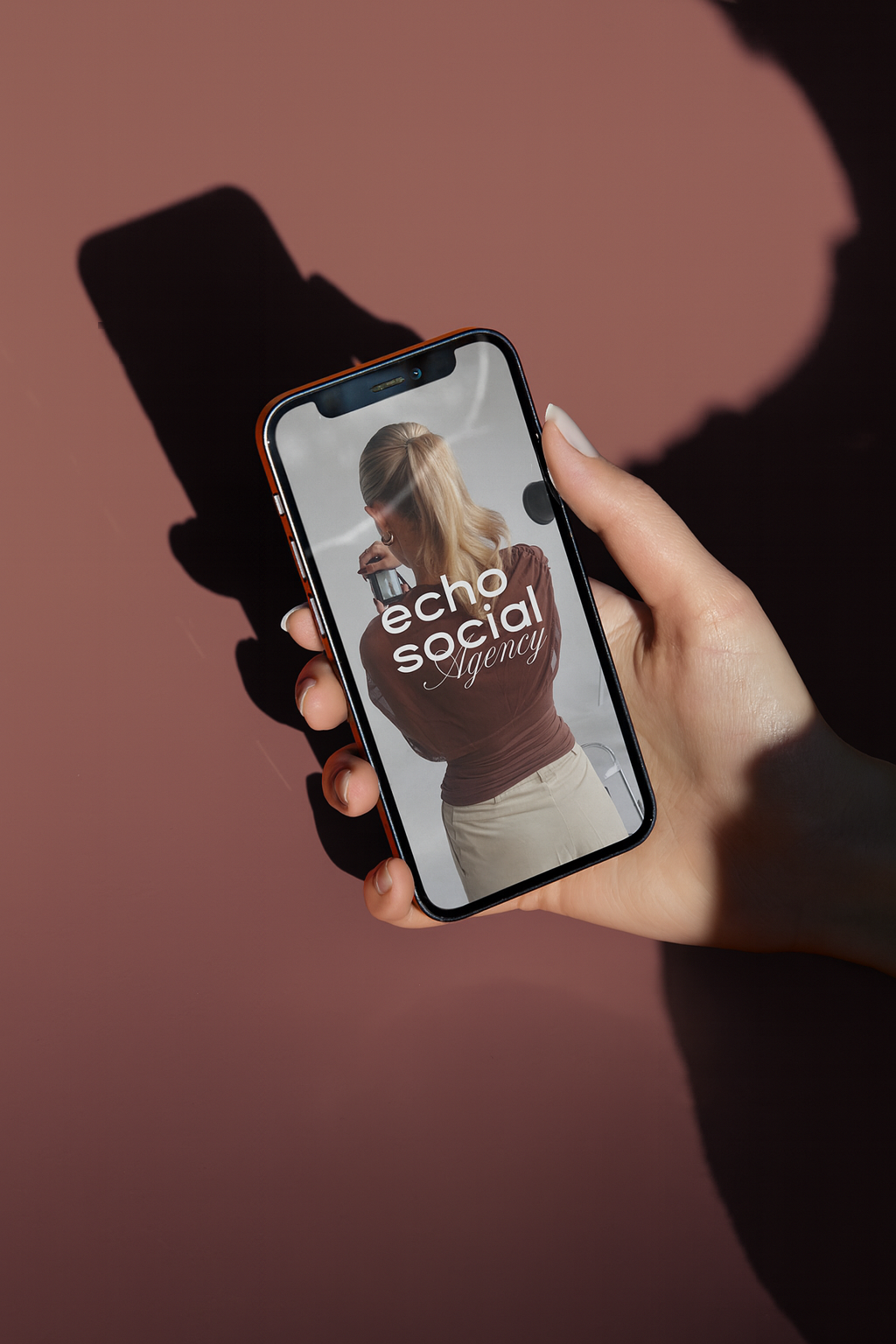 A person holding a smartphone with a photo of a woman on the screen. The woman is drinking coffee and has blond hair, wearing a brown top and beige pants. The screen displays the text "echo social Agency." The background is pink with a shadow of the phone cast on it.
