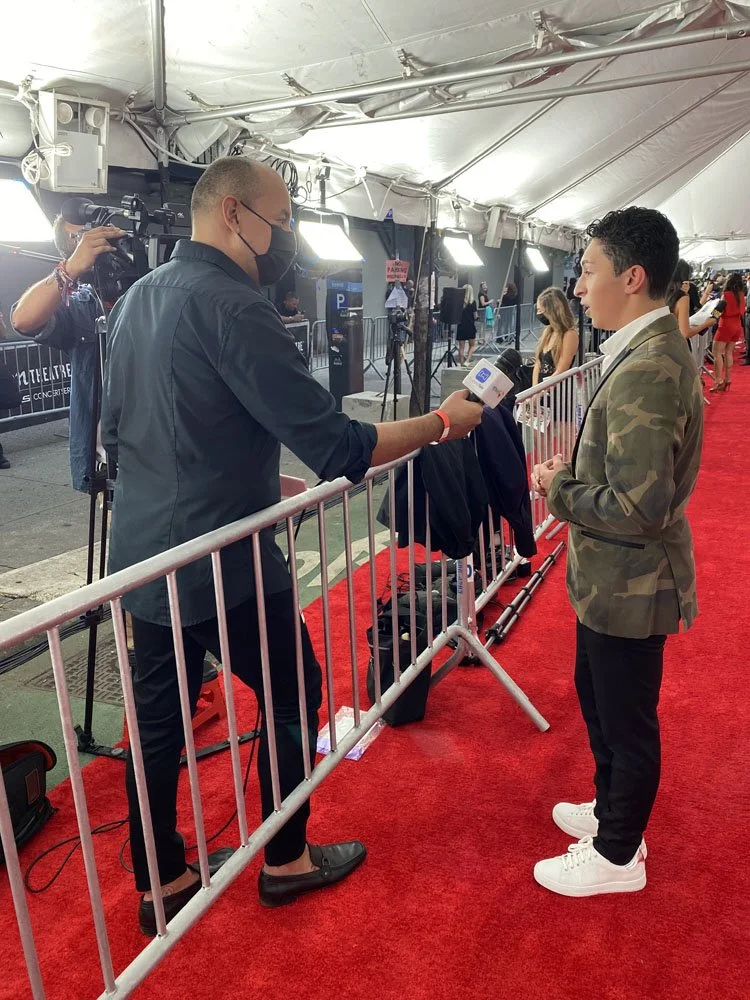 A man in a camouflage blazer and white sneakers speaking to a reporter holding a microphone at an outdoor event on a red carpet, with a security barrier separating them. Several other attendees are visible in the background under a tent.