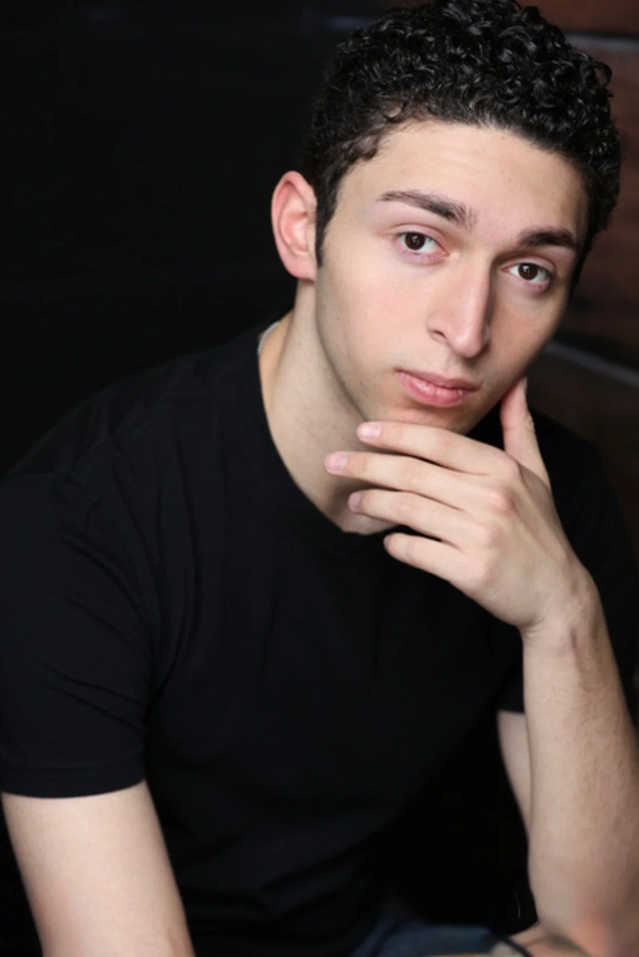 A young man with short, curly dark hair and light skin, wearing a black T-shirt, sitting with his hand on his chin, looking directly at the camera against a dark background.