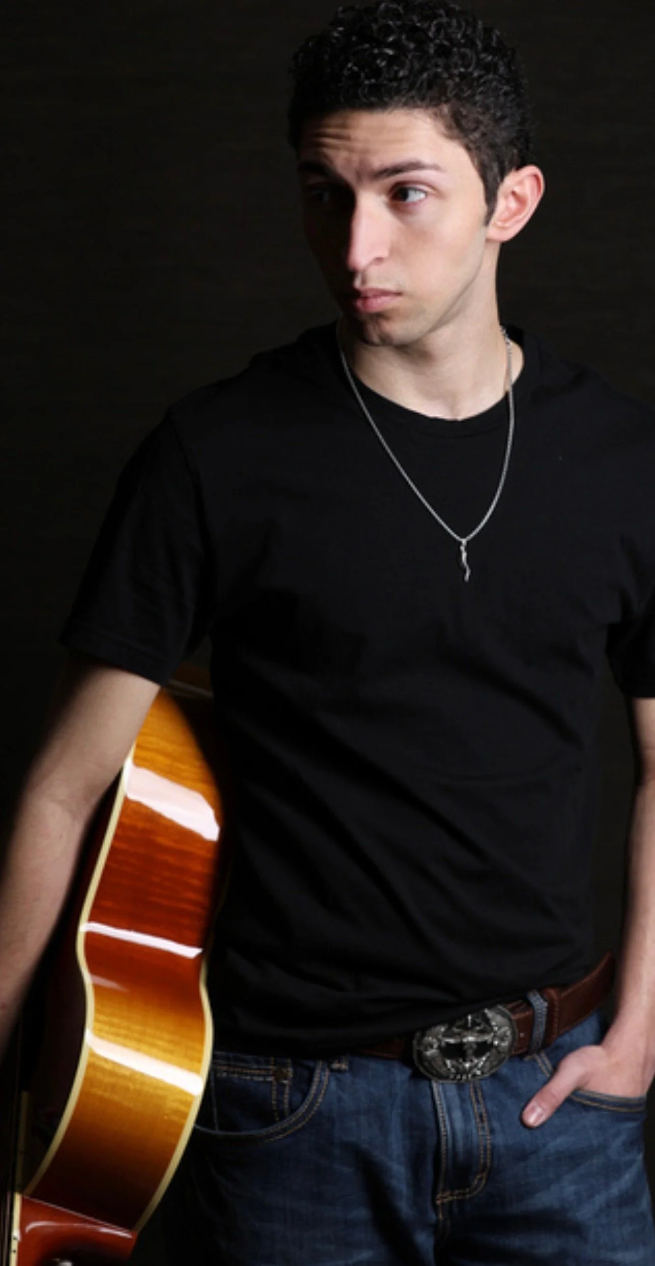 Young man with curly black hair wearing a black T-shirt and a silver chain necklace holding an acoustic guitar with a glossy sunburst finish, standing against a dark background.