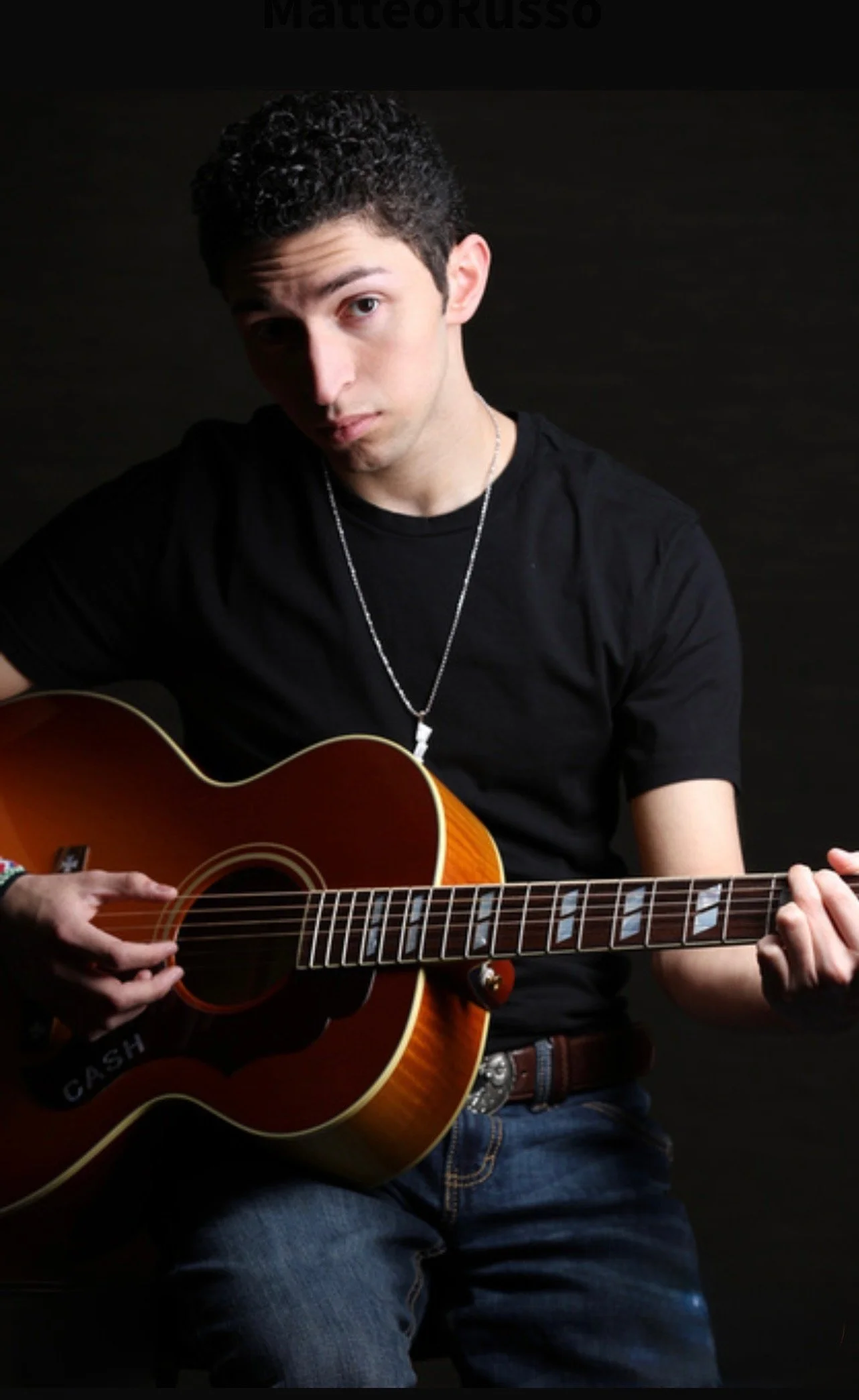 A young man with dark, curly hair playing an acoustic guitar, wearing a black T-shirt, jeans, and a silver chain, against a dark background.