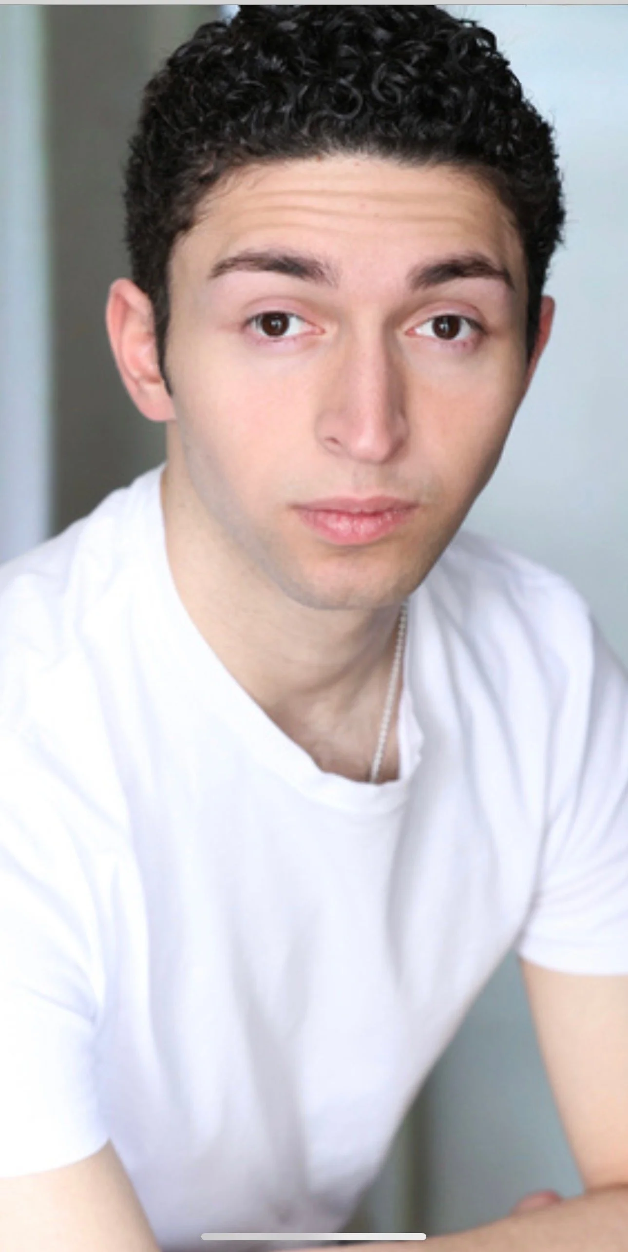 A young man with curly dark hair wearing a white t-shirt and a silver chain, looking slightly to the side with a neutral expression.