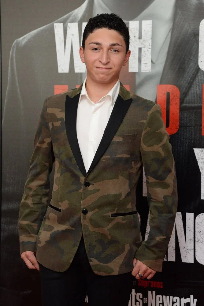 A young man with short dark curly hair standing in front of a promotional backdrop. He is wearing a white shirt and a camo-patterned blazer with black trim. He is smiling slightly and has his hands at his sides.