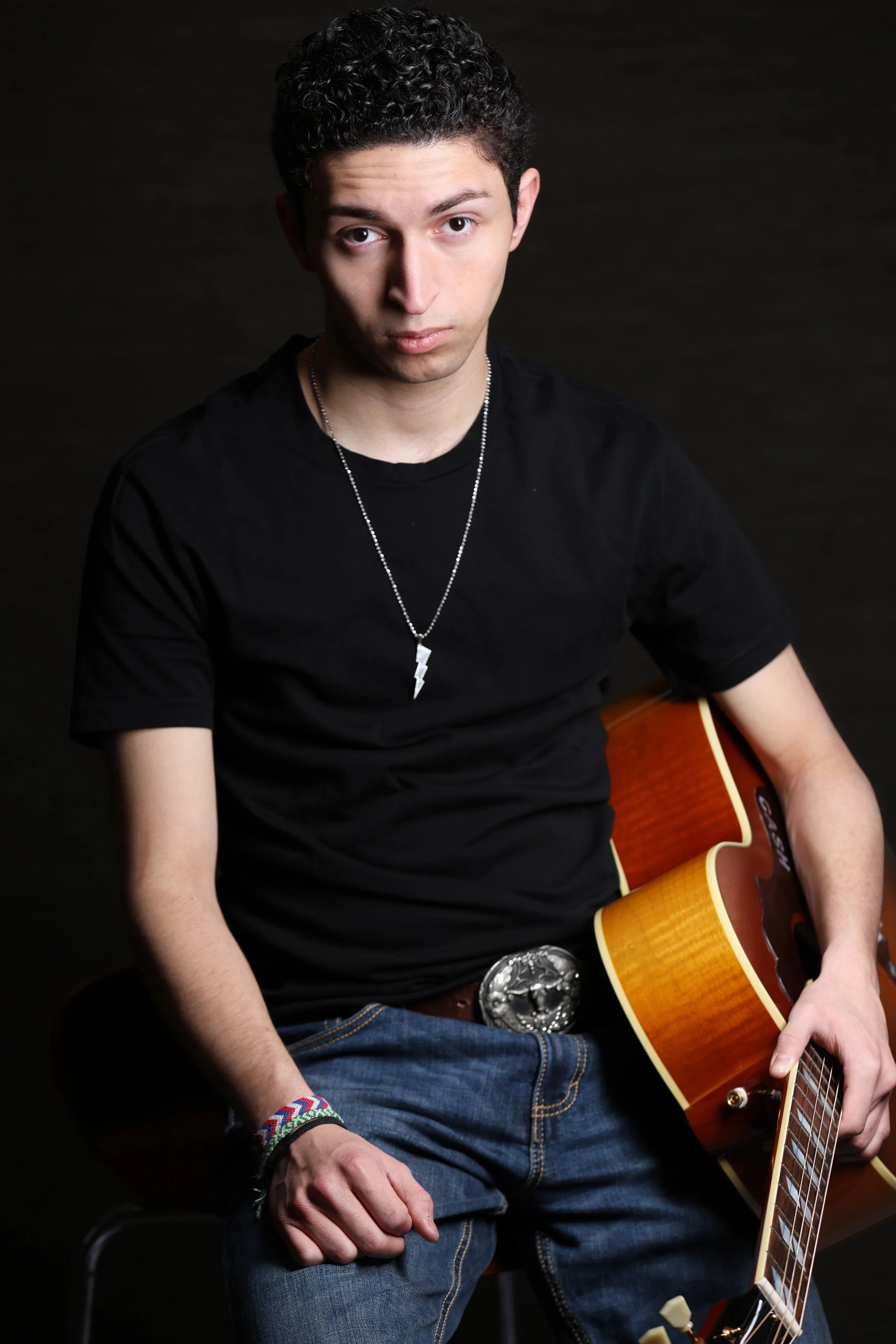 A young man with curly dark hair, wearing a black T-shirt, blue jeans, and a large belt buckle, holding an acoustic guitar, and wearing a lightning bolt necklace, sitting against a dark background.