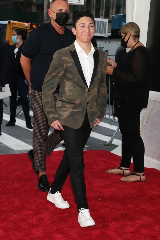 Young man in a white shirt, camouflage blazer, black pants, and white sneakers walking on a red carpet, with two adults in masks in the background at an indoor event.