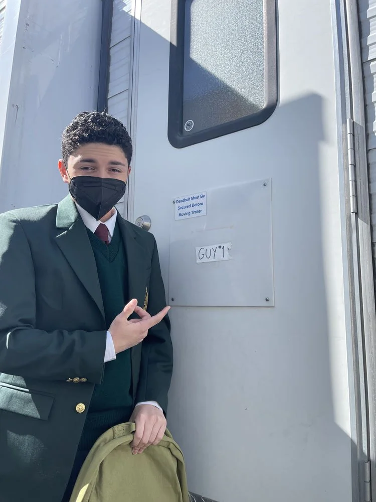 Young man in school uniform and face mask pointing at a sign on a trailer that says 'GUY*1'.