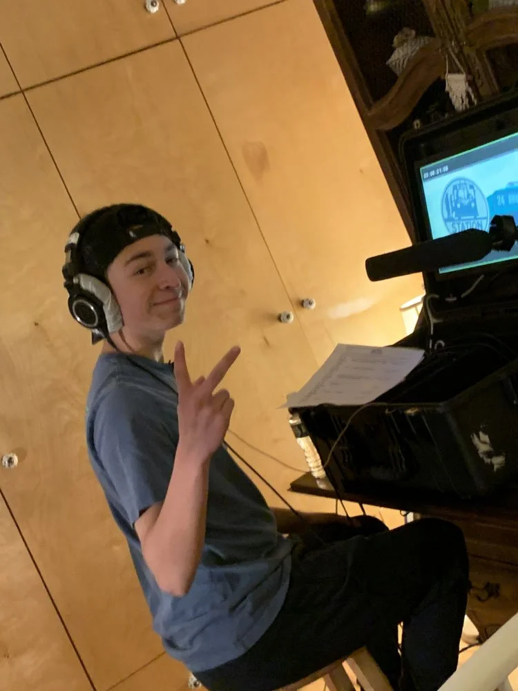 Young man sitting at a piano, wearing headphones, holding sheet music, and making a peace sign with his hand.