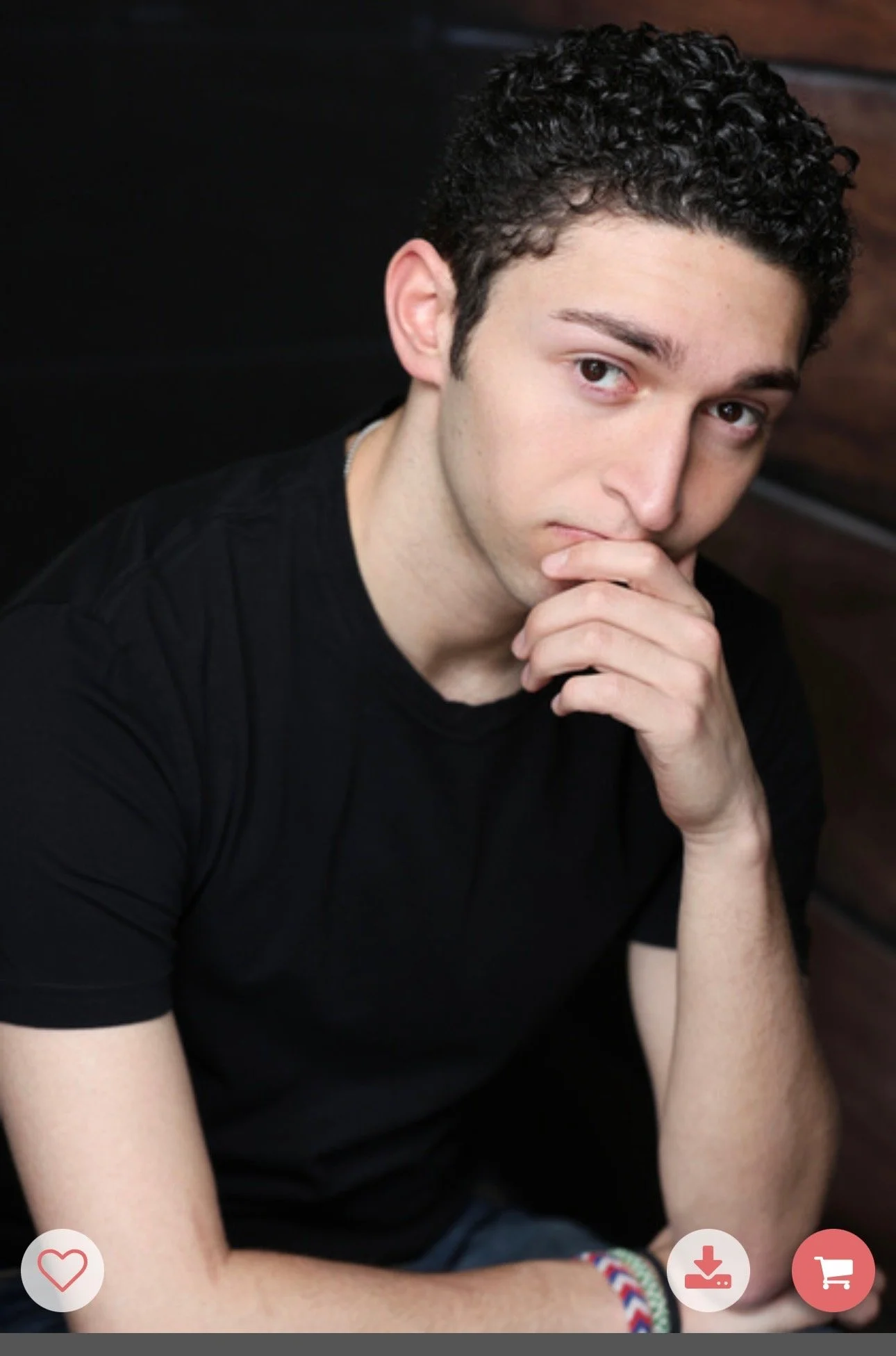 A young man with dark curly hair, wearing a black t-shirt, sitting against a black background with wooden paneling, hand covering his mouth, looking at the camera.
