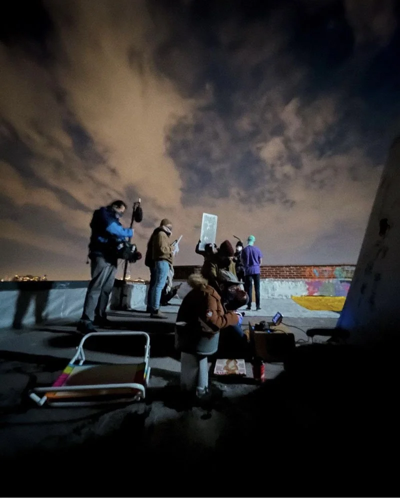 People gathered on a rooftop at night, some taking photos or holding signs, with a cloudy sky overhead and city lights in the distance.